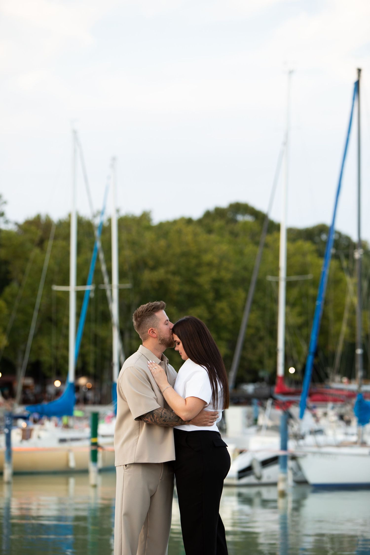 Romantic couple embracing at a marina with sailboats in the background on a cloudy day.