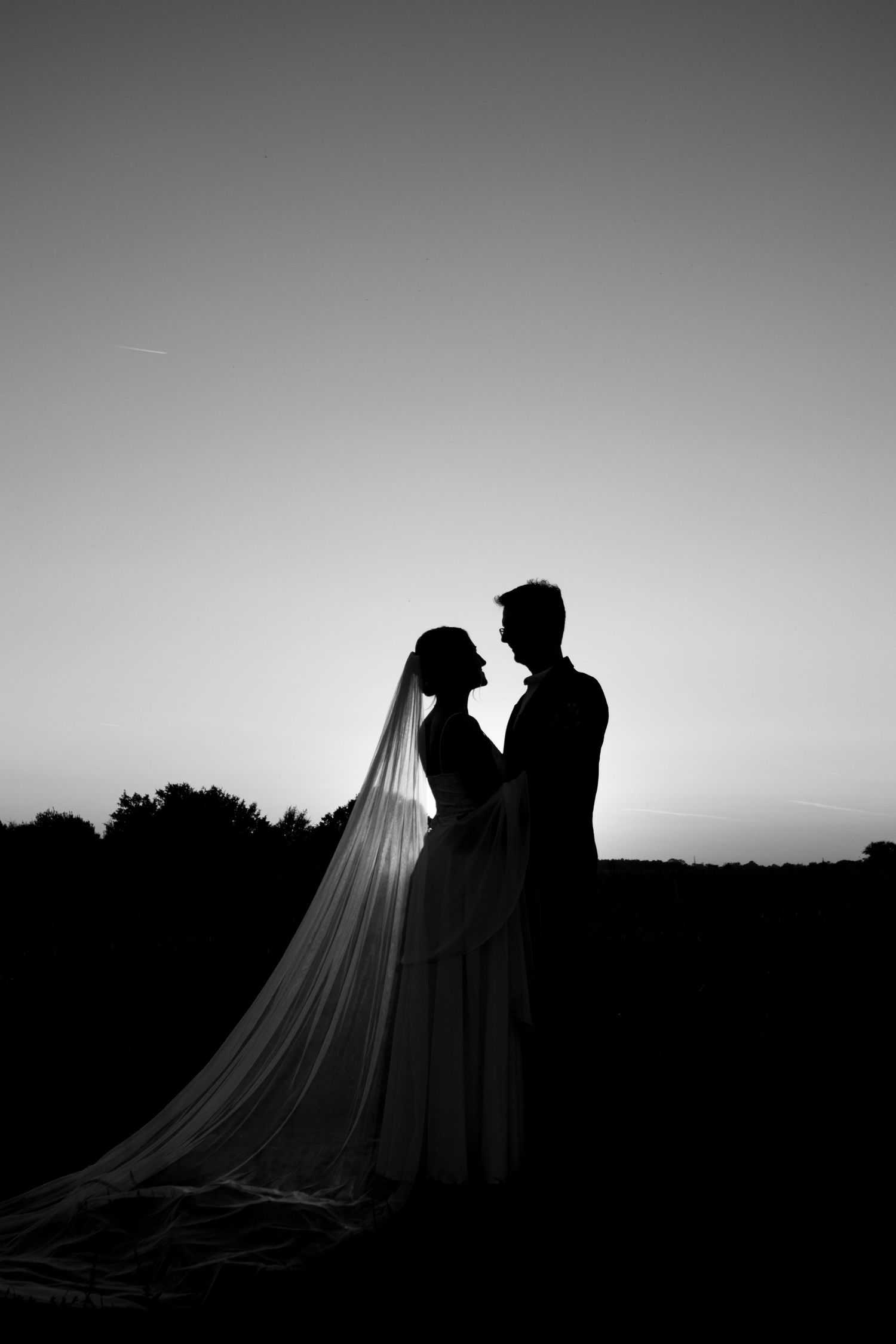 Romantic silhouette of a wedding couple against a sunset sky with flowing veil creating a dramatic outline.