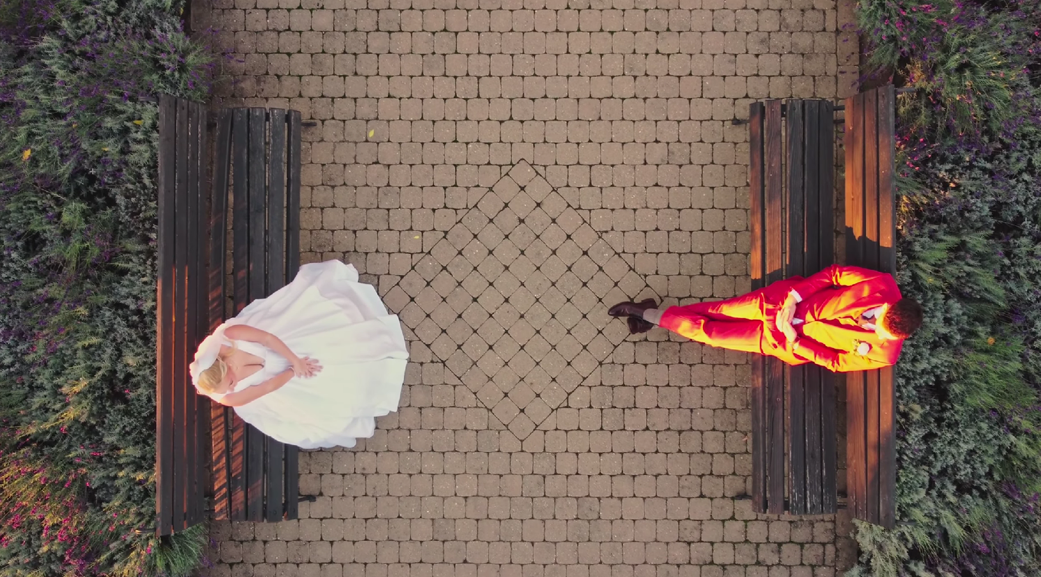 Aerial view of bride in white dress and groom in red suit sitting on benches on a brick path.