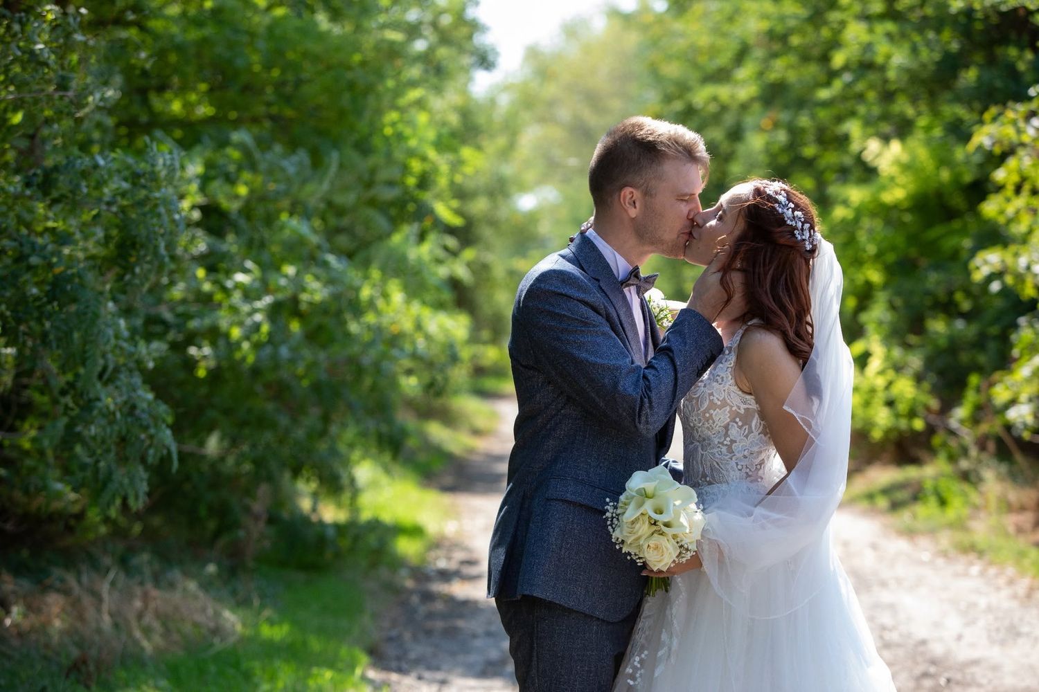 A bride and groom share a romantic kiss outdoors on a garden path surrounded by lush green trees.