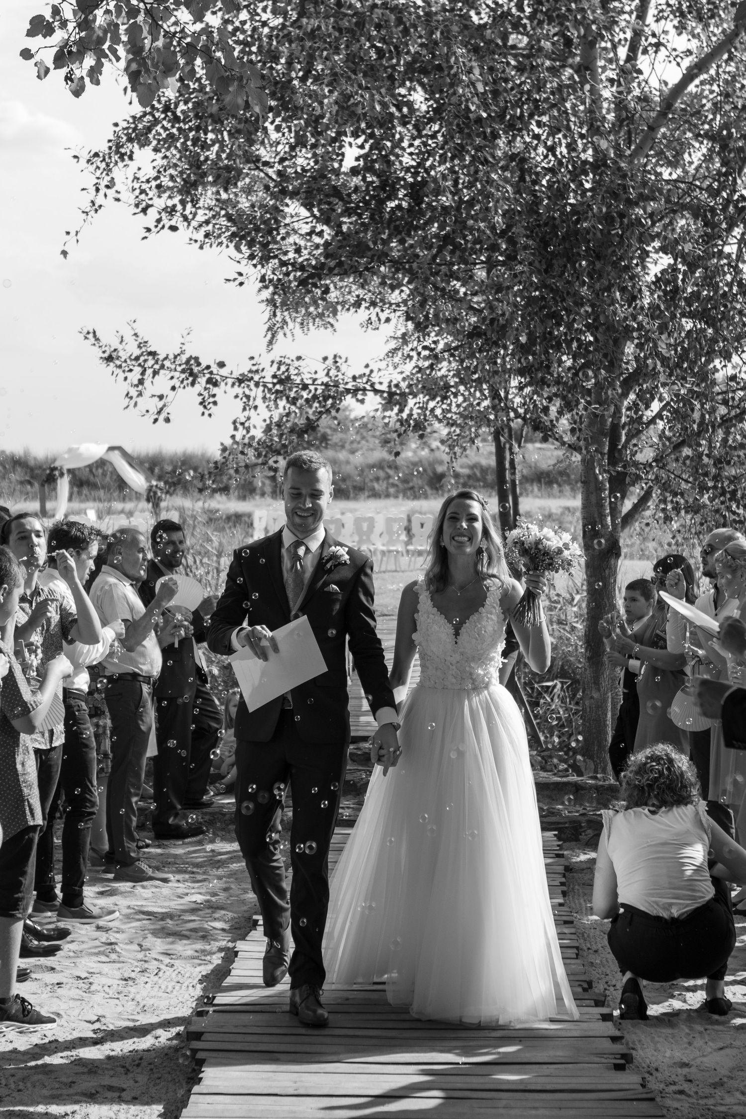 Wedding celebration with guests throwing confetti under shaded trees.