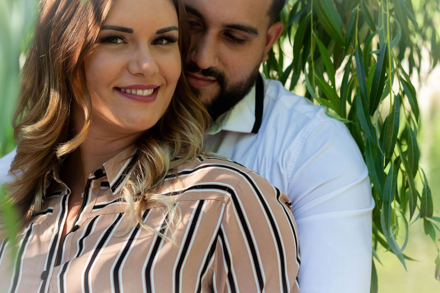 Close up of couple embracing outdoors with woman wearing striped blouse against green foliage.