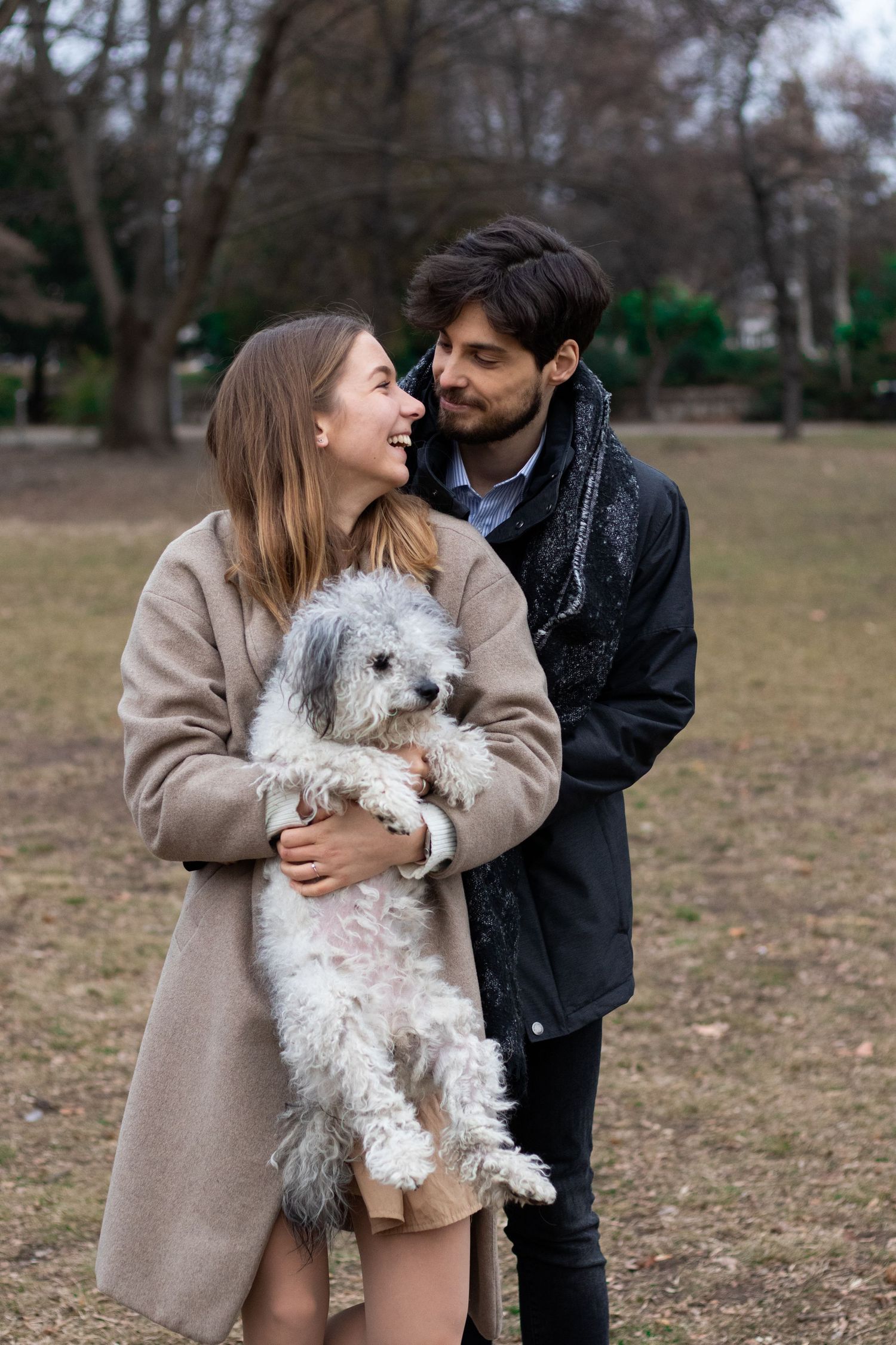 Couple holding small white dog while sharing intimate moment in winter park setting.