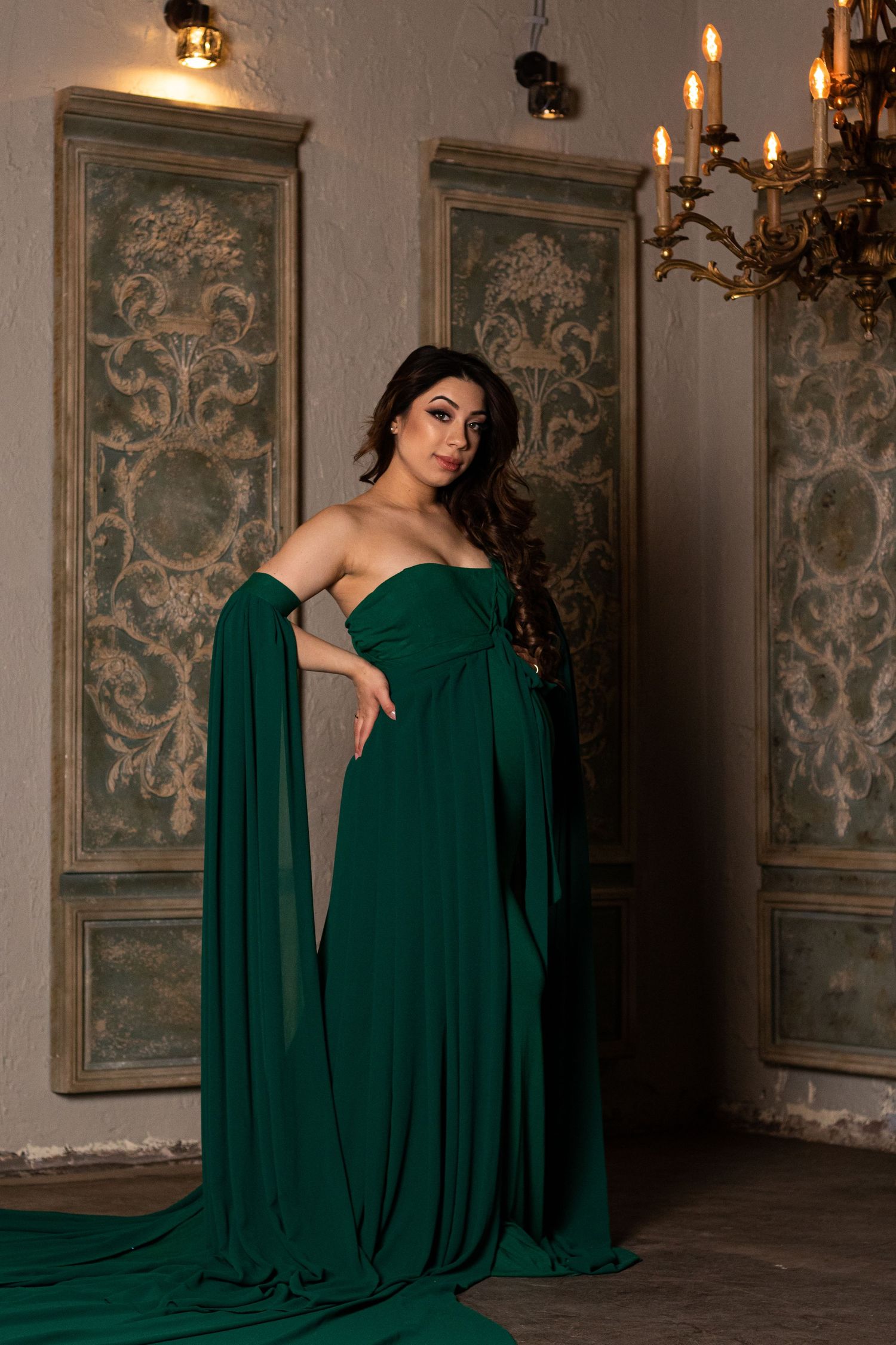 Dramatic maternity portraits with flowing emerald gown against vintage wall art and chandeliers.