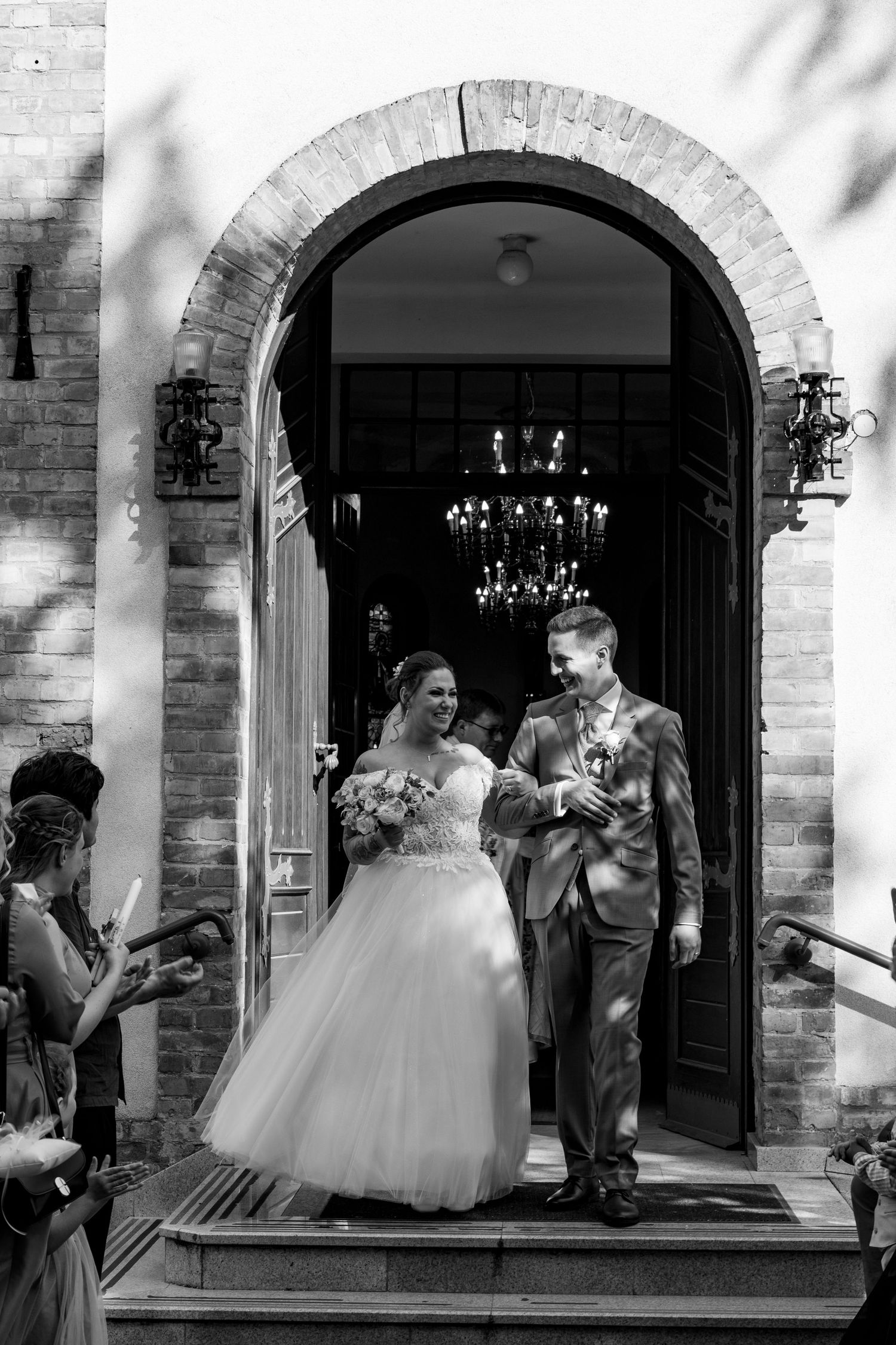 A wedding couple exits through a brick archway with elegant chandeliers visible in the background.