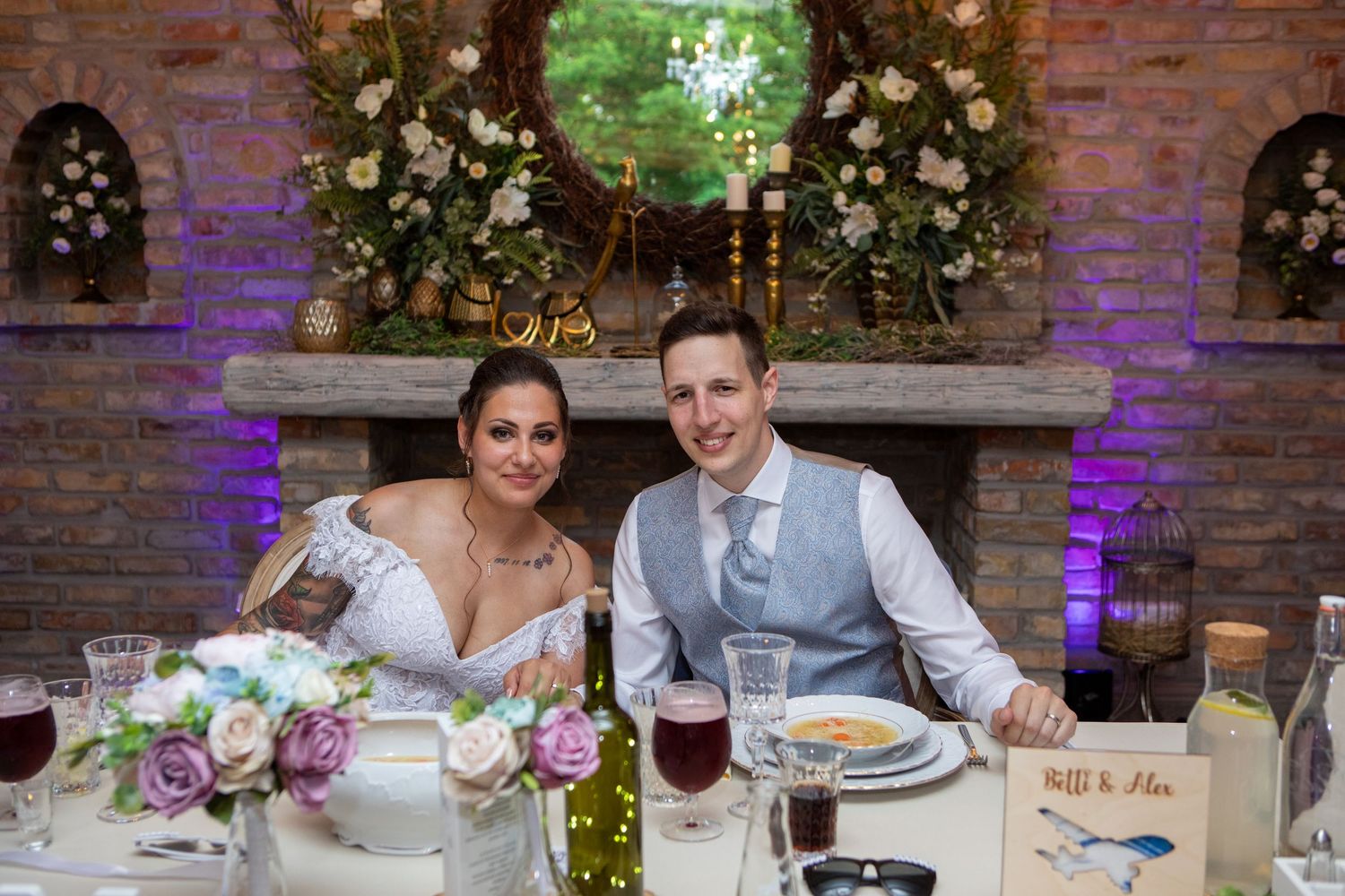 Couple sits at decorated sweetheart table with floral arch and purple lighting.