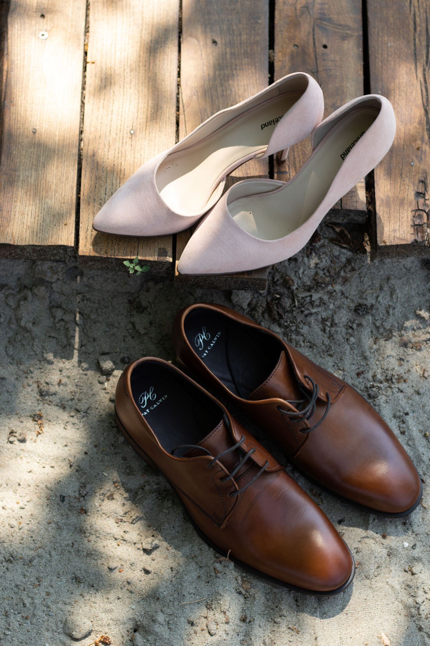 Brown leather dress shoes paired with white bridal heels on rustic wooden deck.