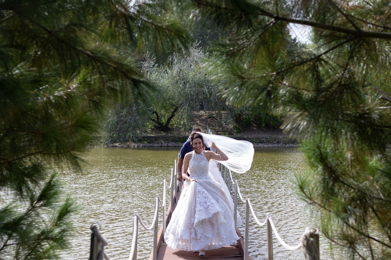 A flowing white wedding dress billows in the wind on a wooden dock over a serene lake.