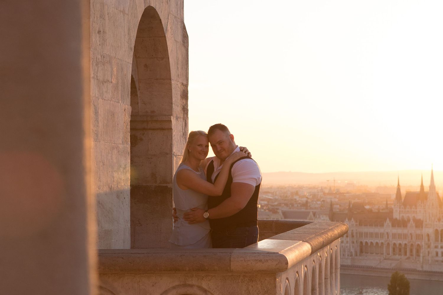Romantic sunset moment of couple embracing on historic stone balcony overlooking city.