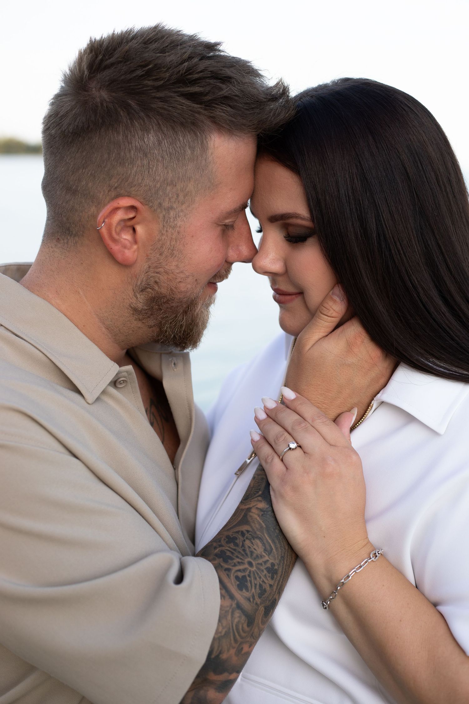 Romantic couple touching foreheads tenderly outdoors near water, woman wearing engagement ring and bracelet.