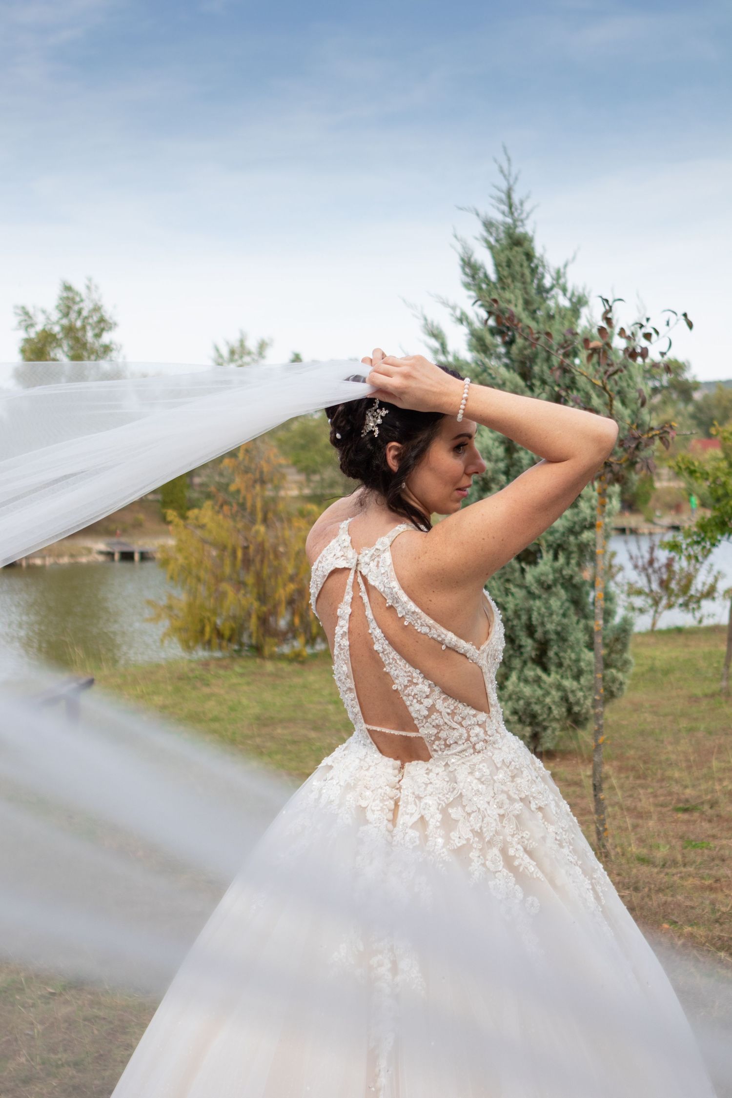 A delicate lace wedding dress with flowing veil captures movement against a natural lakeside backdrop.