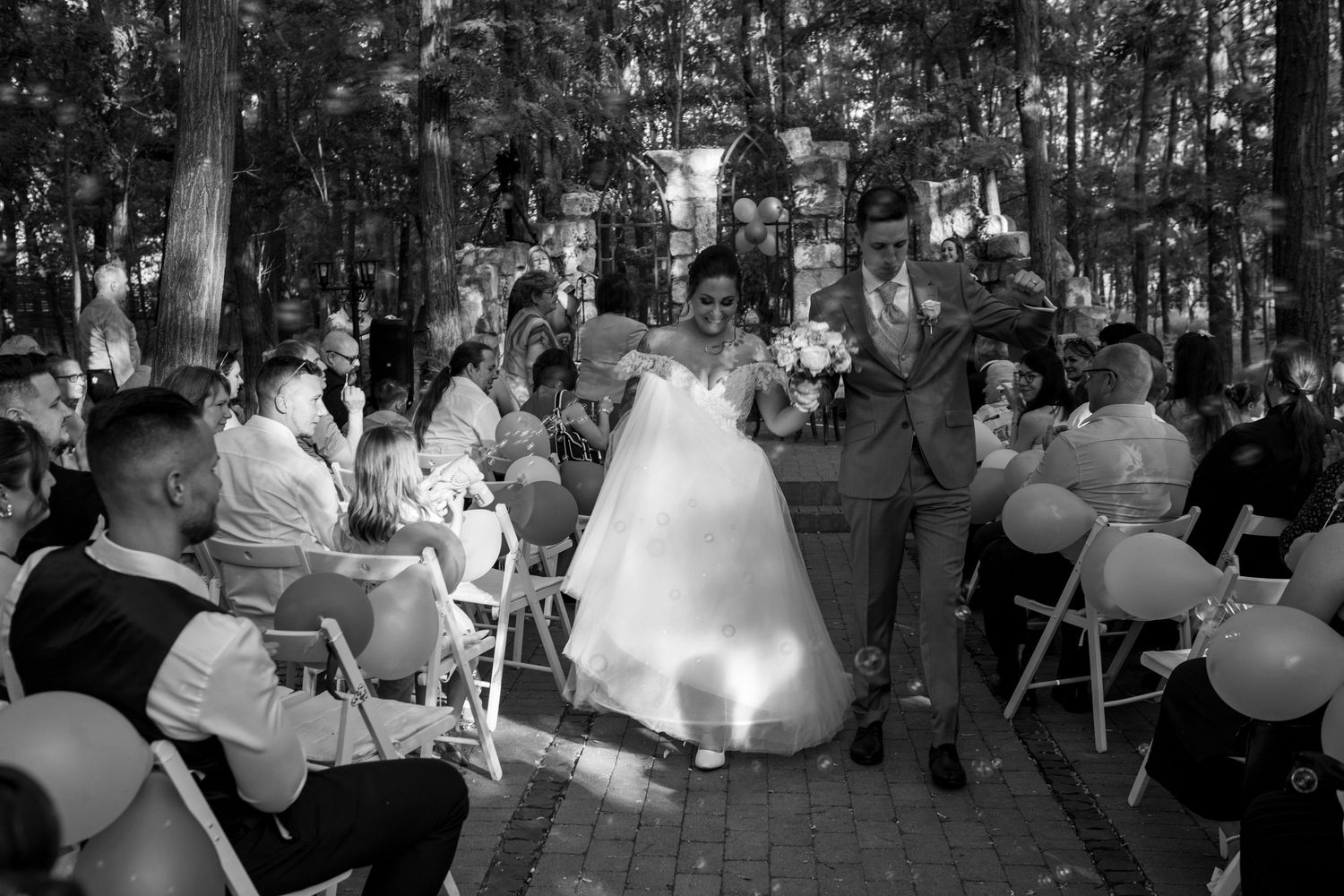 Black and white photo of outdoor wedding ceremony in wooded setting with guests seated on folding chairs.