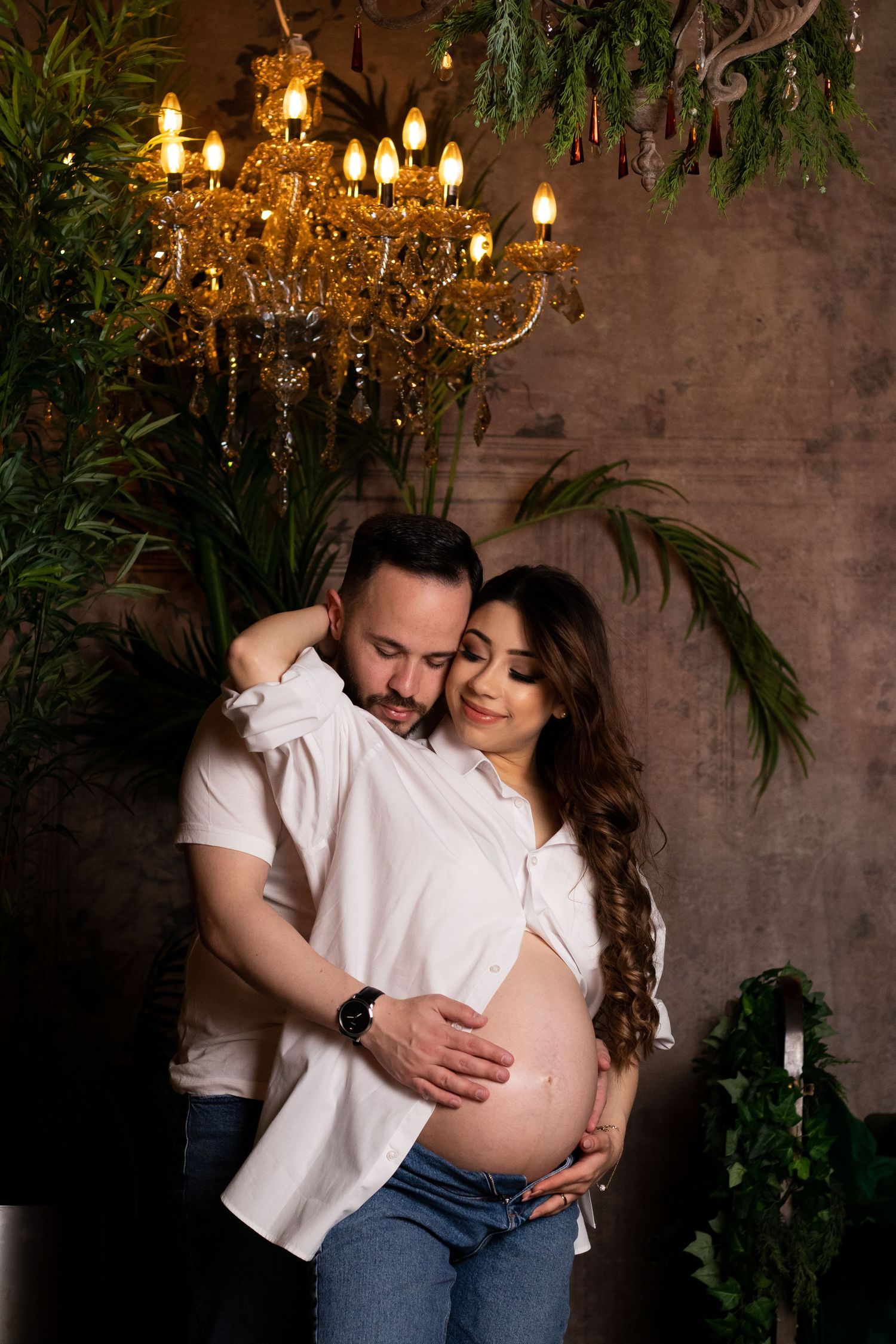 Intimate maternity photoshoot with couple embracing in romantic setting with chandelier and greenery.