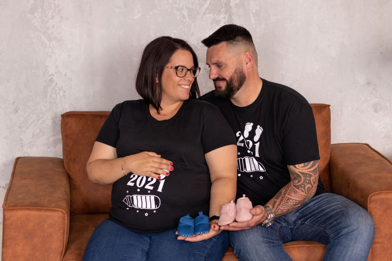 Couple shares tender moment on brown leather couch during indoor maternity session.