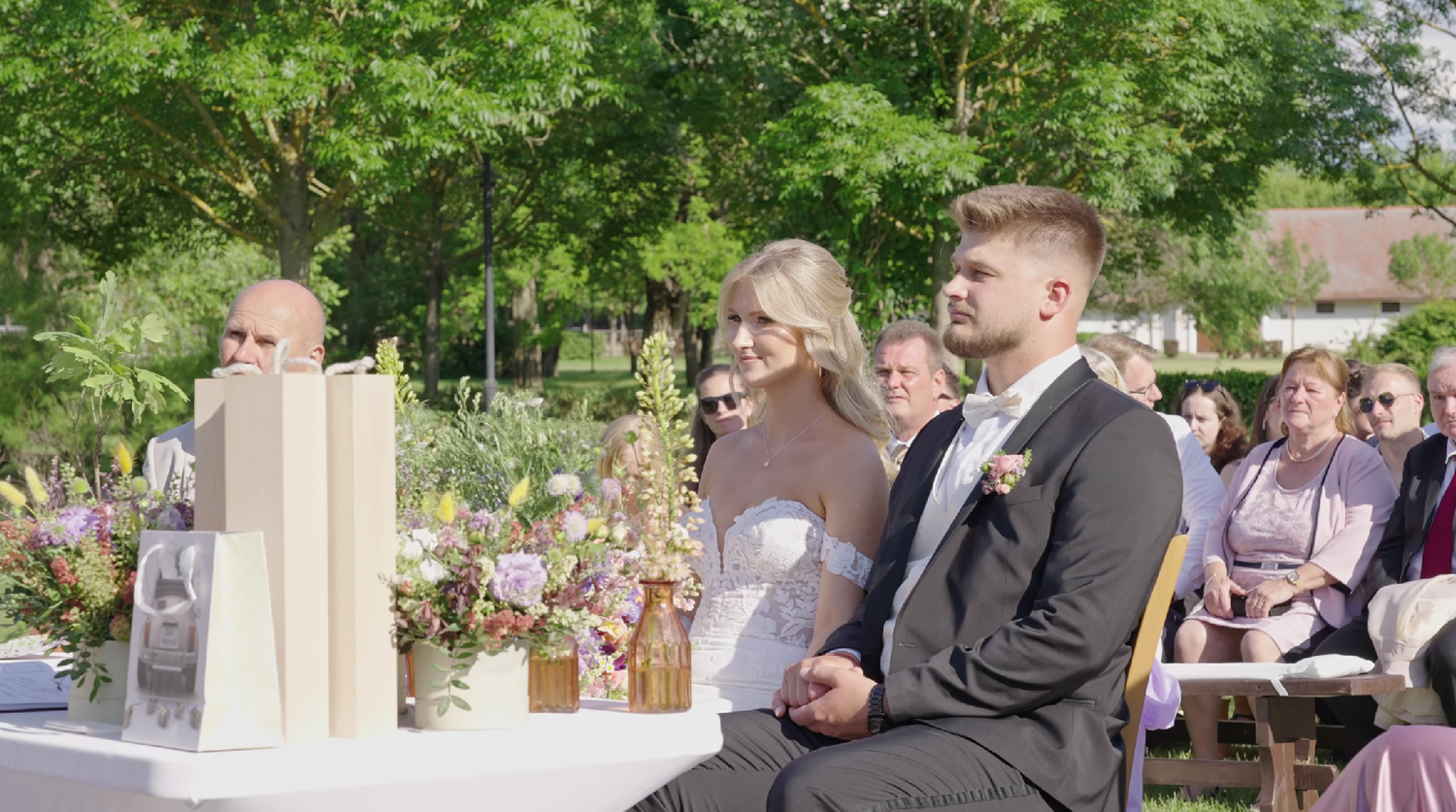 Outdoor wedding ceremony with bride and groom seated, surrounded by floral arrangements and guests in a lush garden setting.