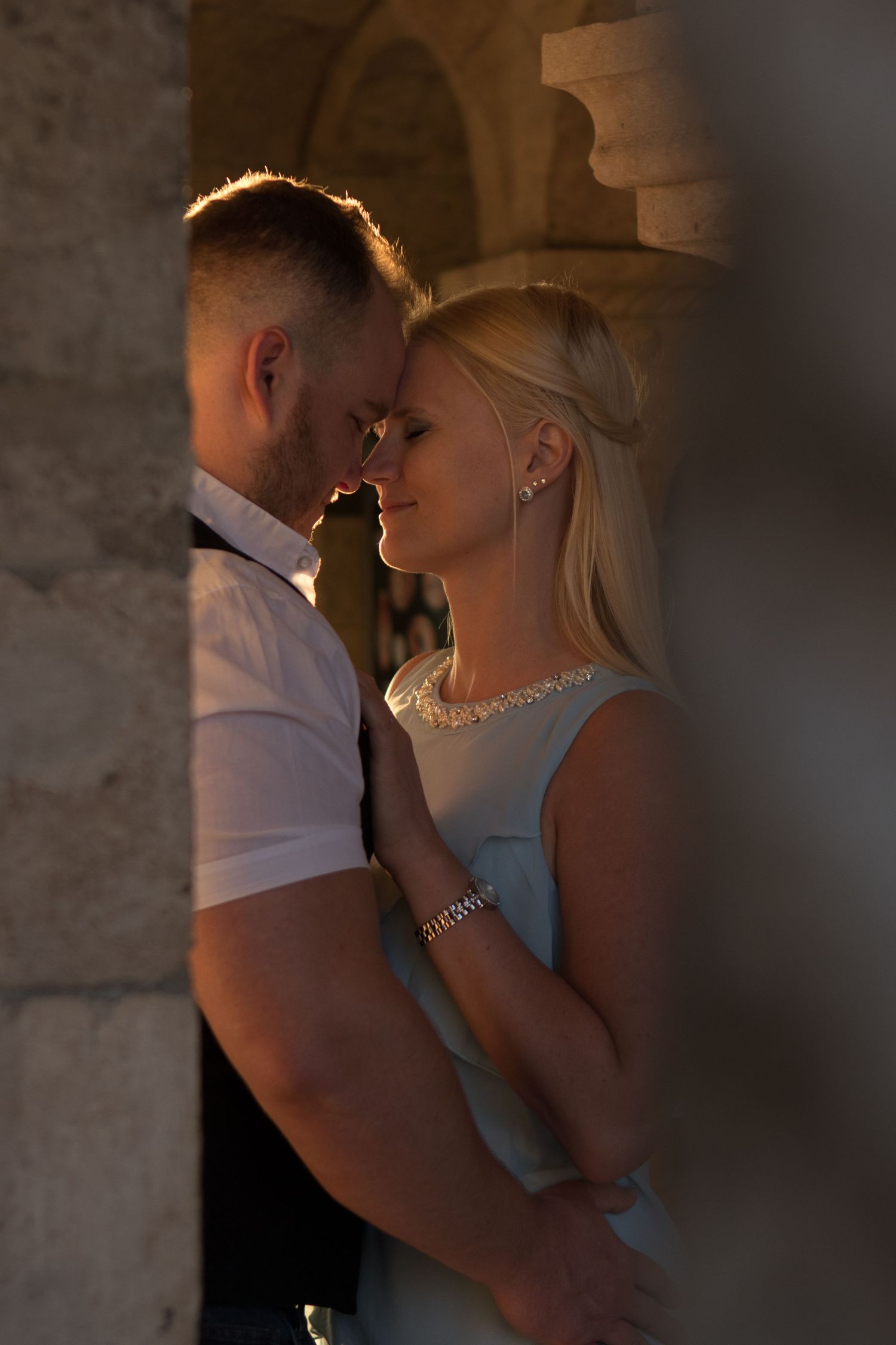Intimate backlit portrait of couple in formal attire sharing tender moment in stone archway.