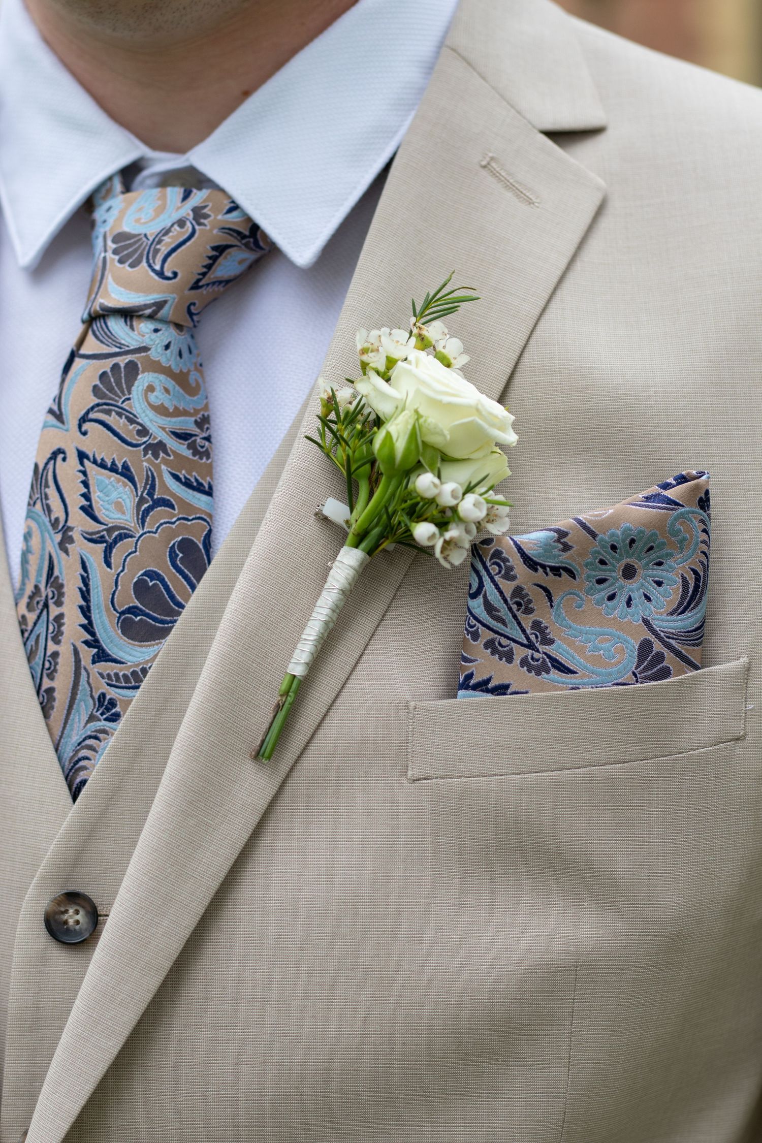 A tan suit jacket with blue paisley tie and white boutonniere.