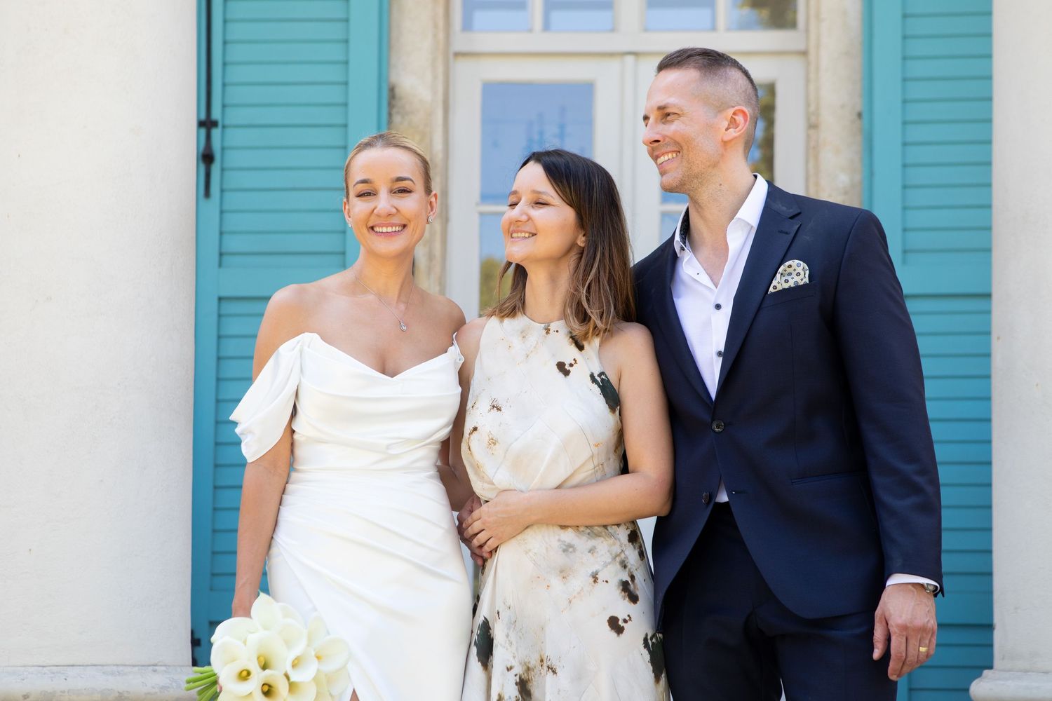 Wedding group photo session against a white building with bright turquoise shutters.