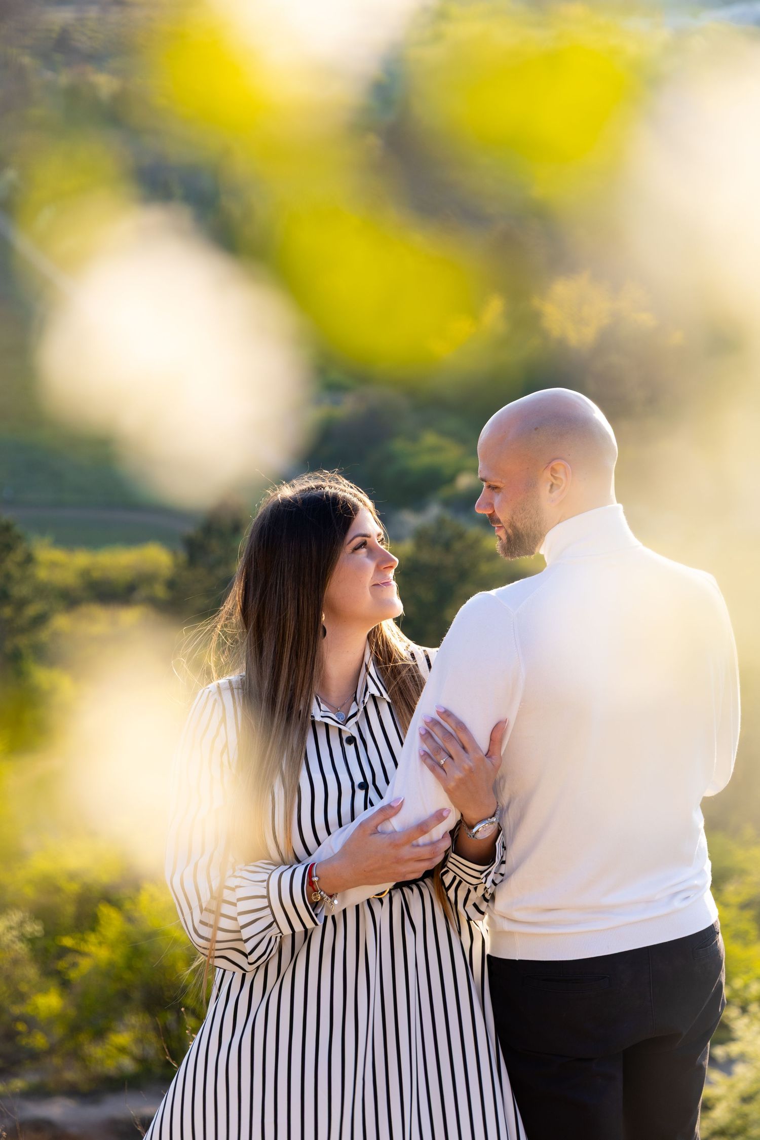 Couple embracing outdoors in golden sunlight, woman in striped dress smiling at bald man in white sweater with green hills behind.
