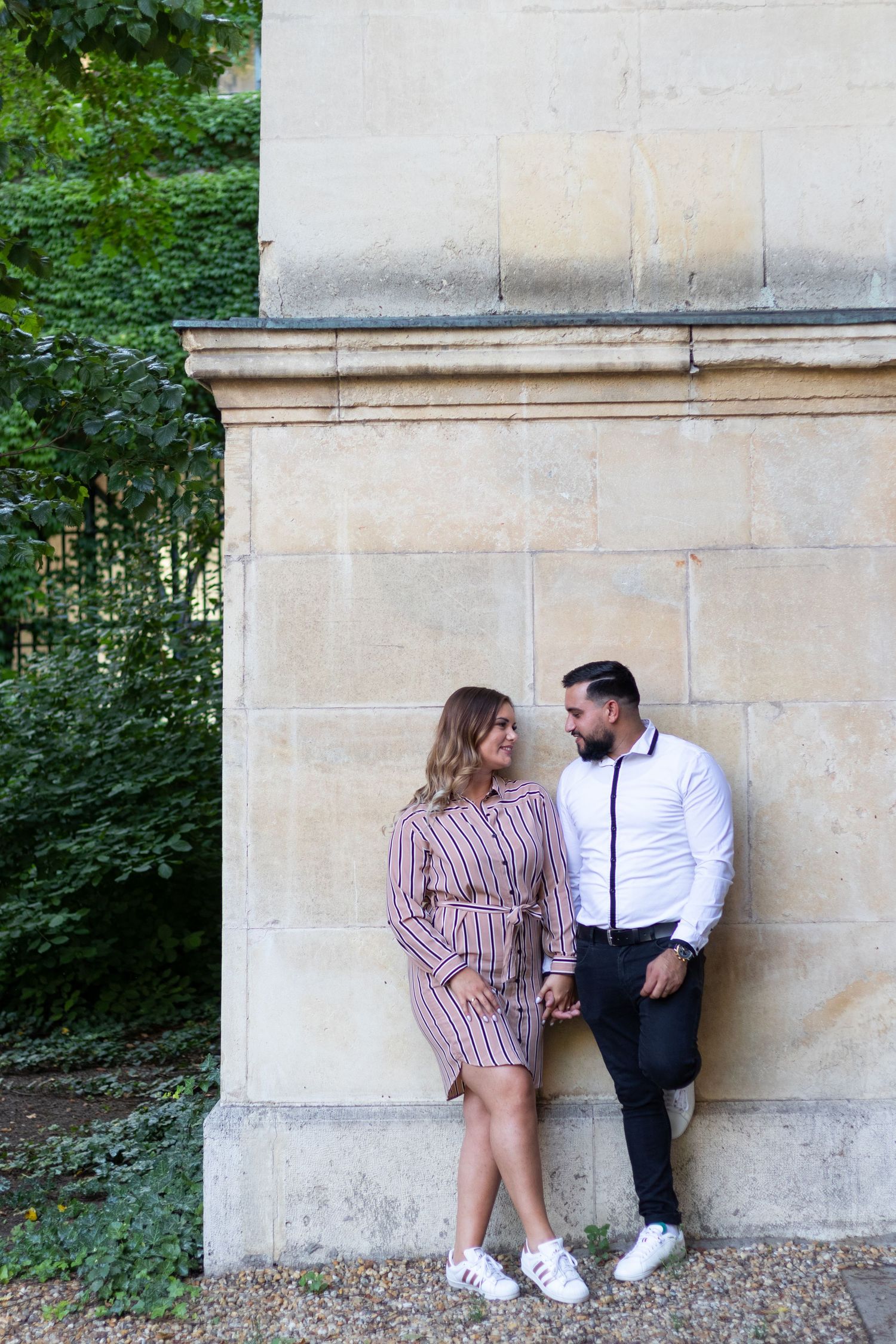 Couple in contrasting outfits posing playfully against a light stone wall outdoors.