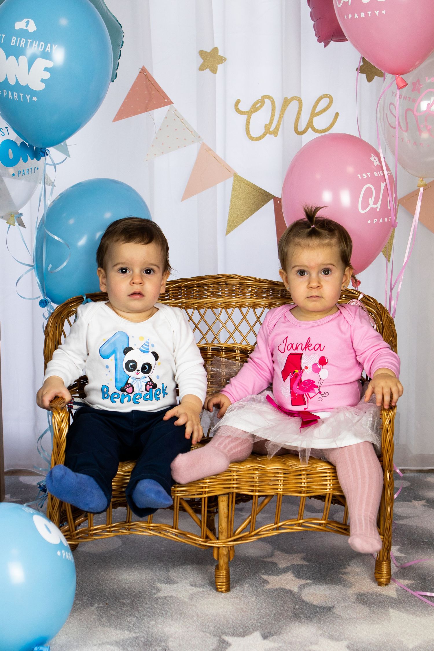 Two babies sitting on wicker bench with birthday balloons and decorative backdrop.