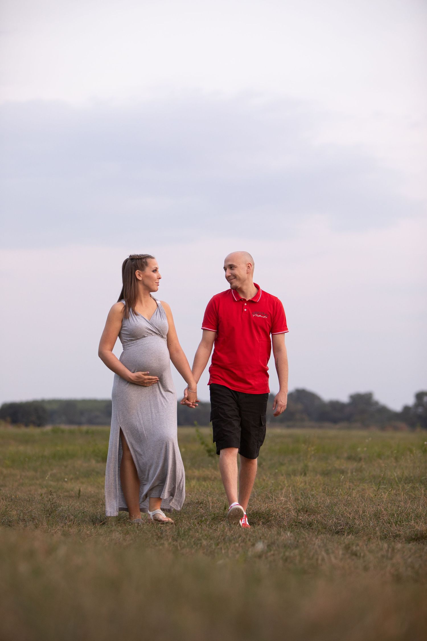 Couple walks hand in hand across an open field during golden hour maternity photoshoot.