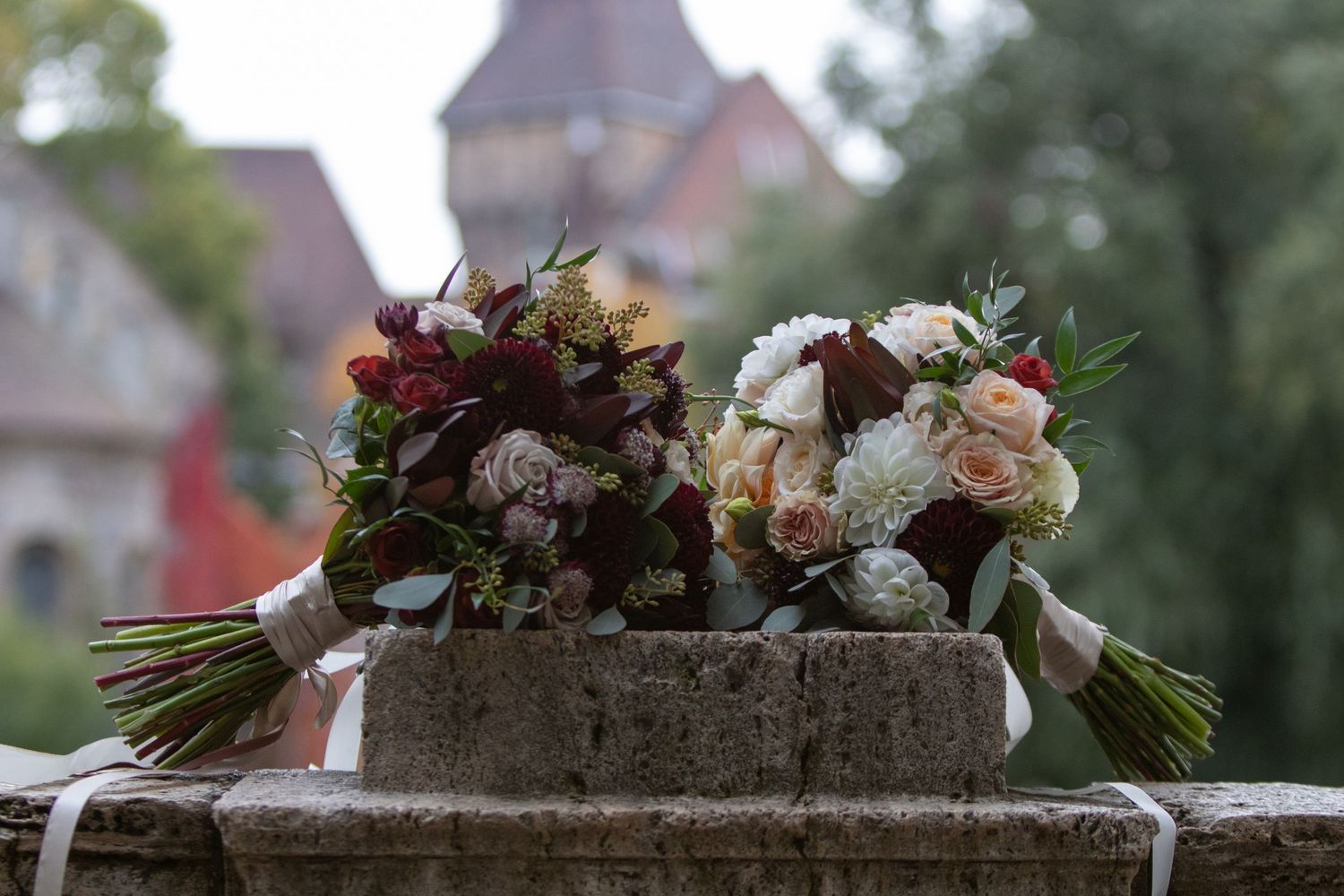 Two elegant wedding bouquets with red and white flowers rest on a stone ledge with a blurred church in the background.