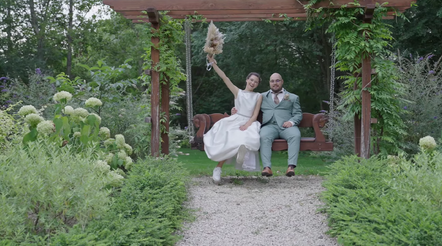 Joyful newlyweds celebrate on a garden bench under a wooden pergola, bride raising bouquet, surrounded by lush greenery.