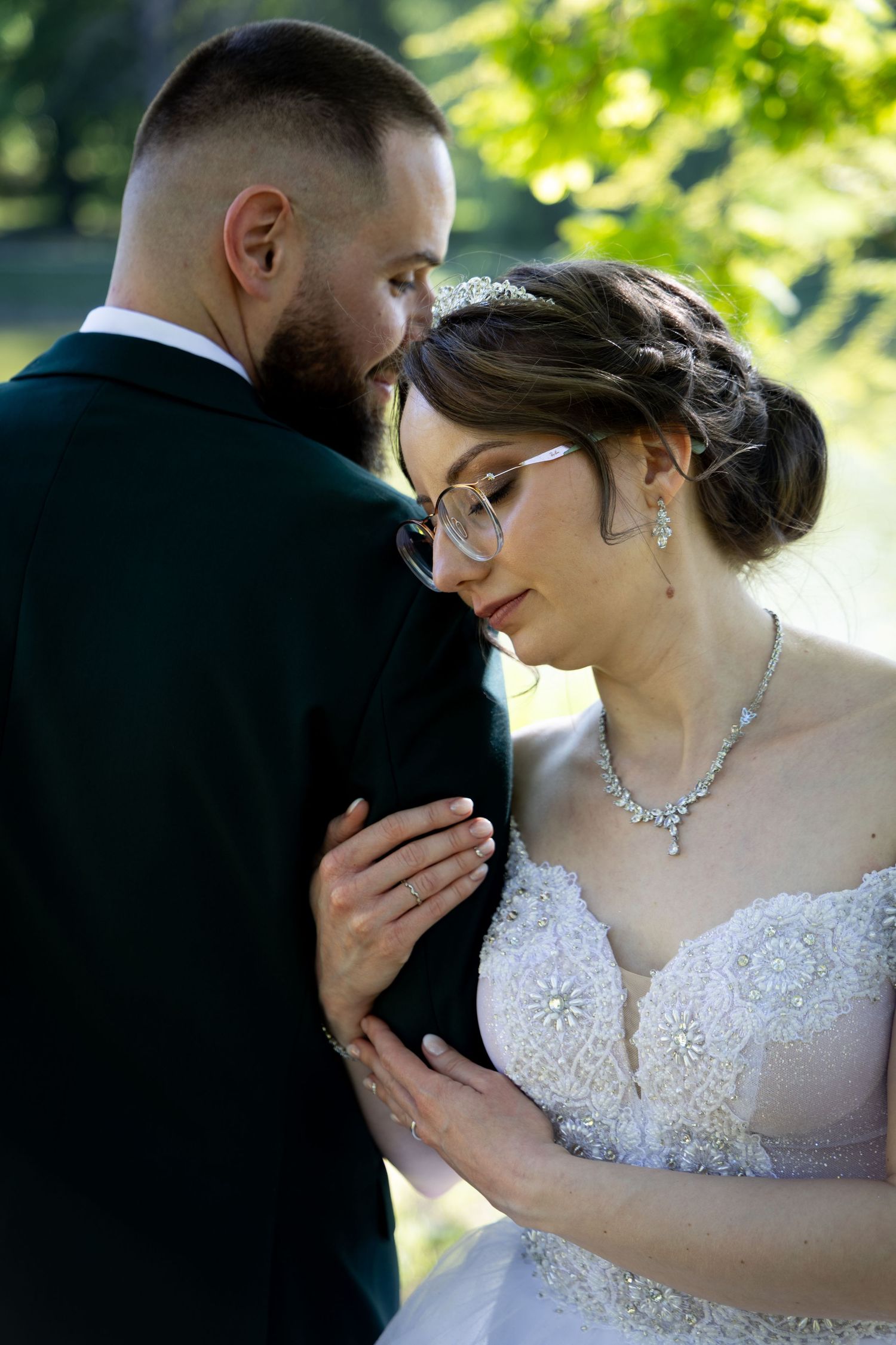 Bride in white lace gown and groom in dark suit share tender moment outdoors, surrounded by lush green trees.