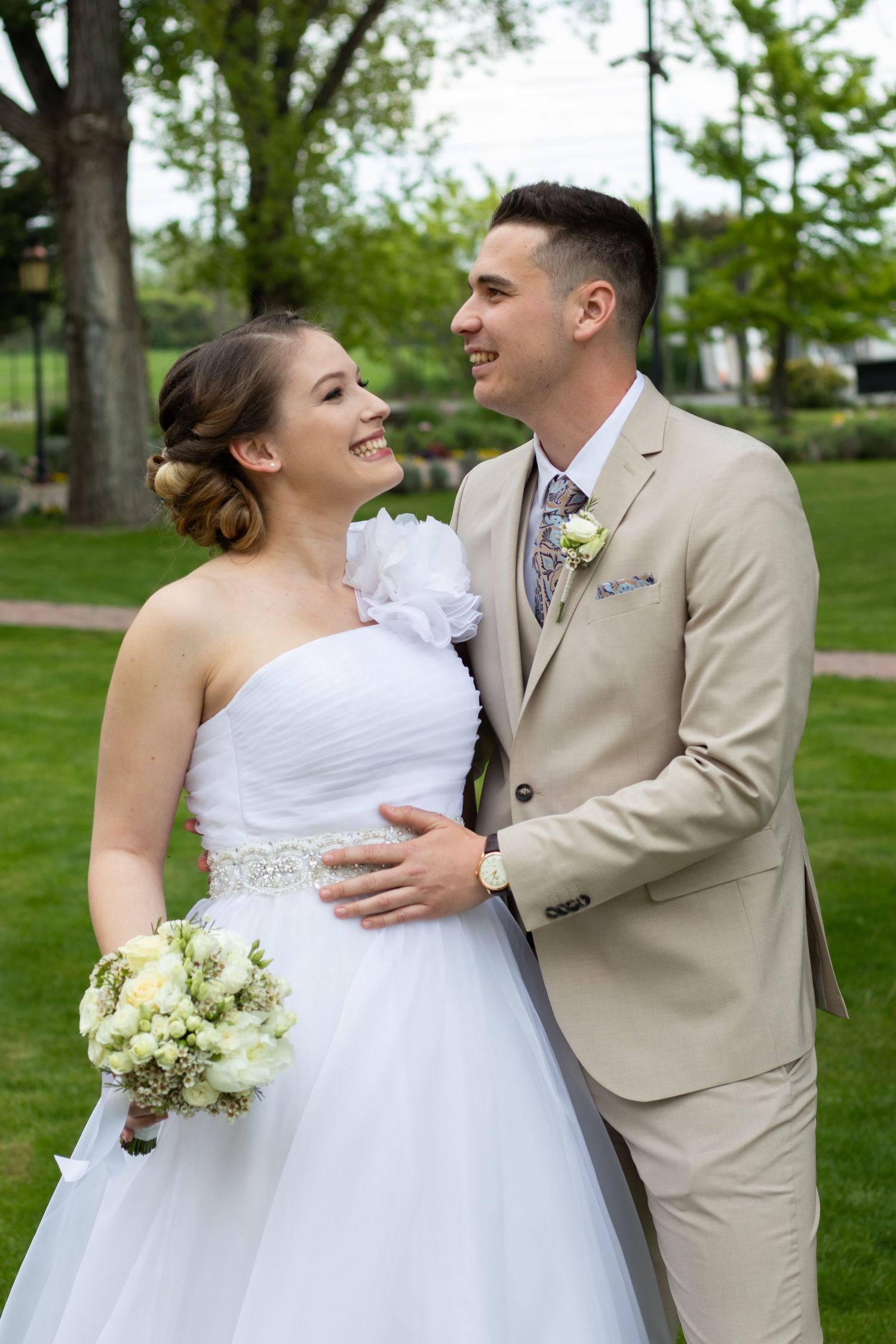 Newlyweds sharing joyful laughter in a park with green trees in the background.