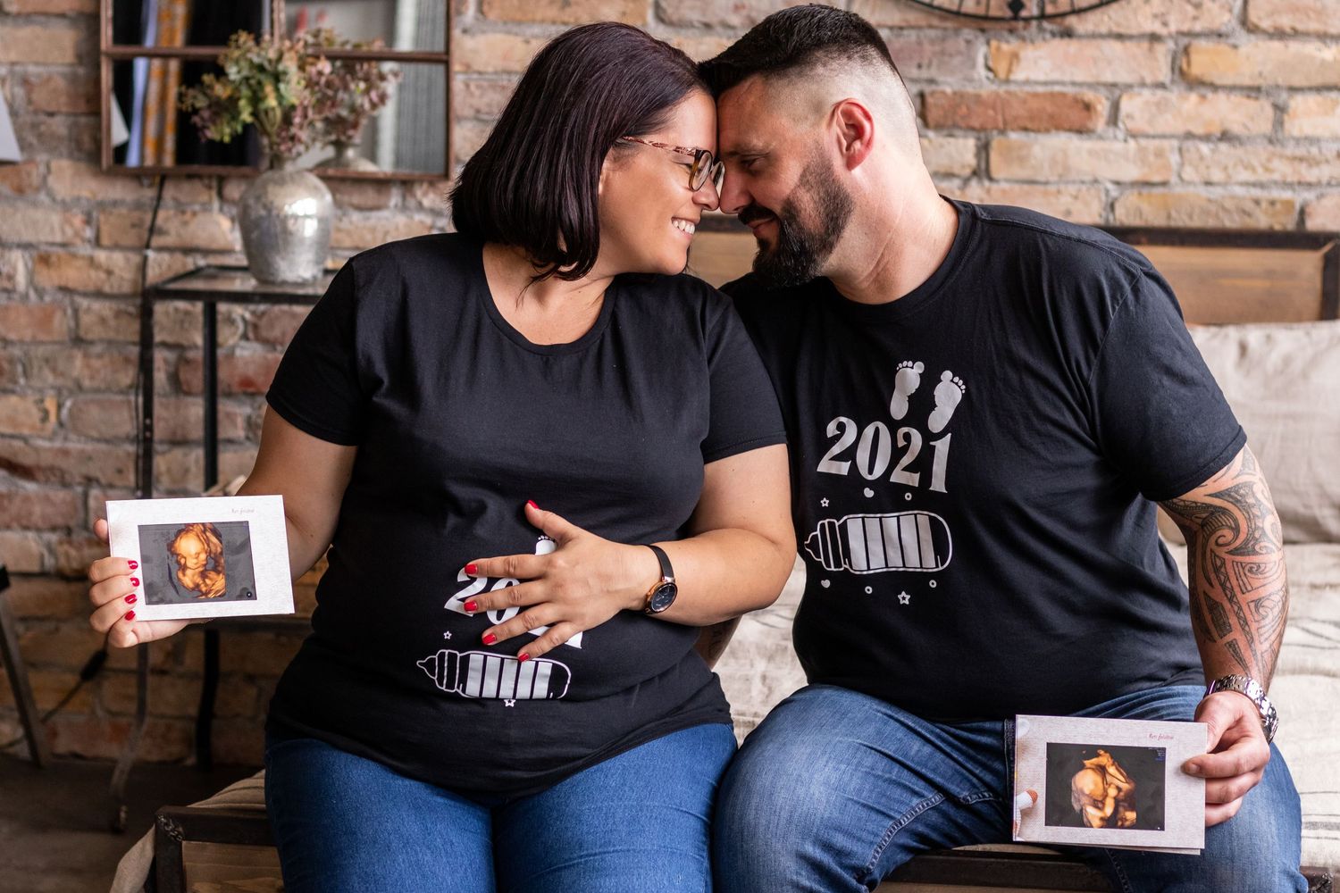 Couple poses together on brown leather couch during indoor maternity session.
