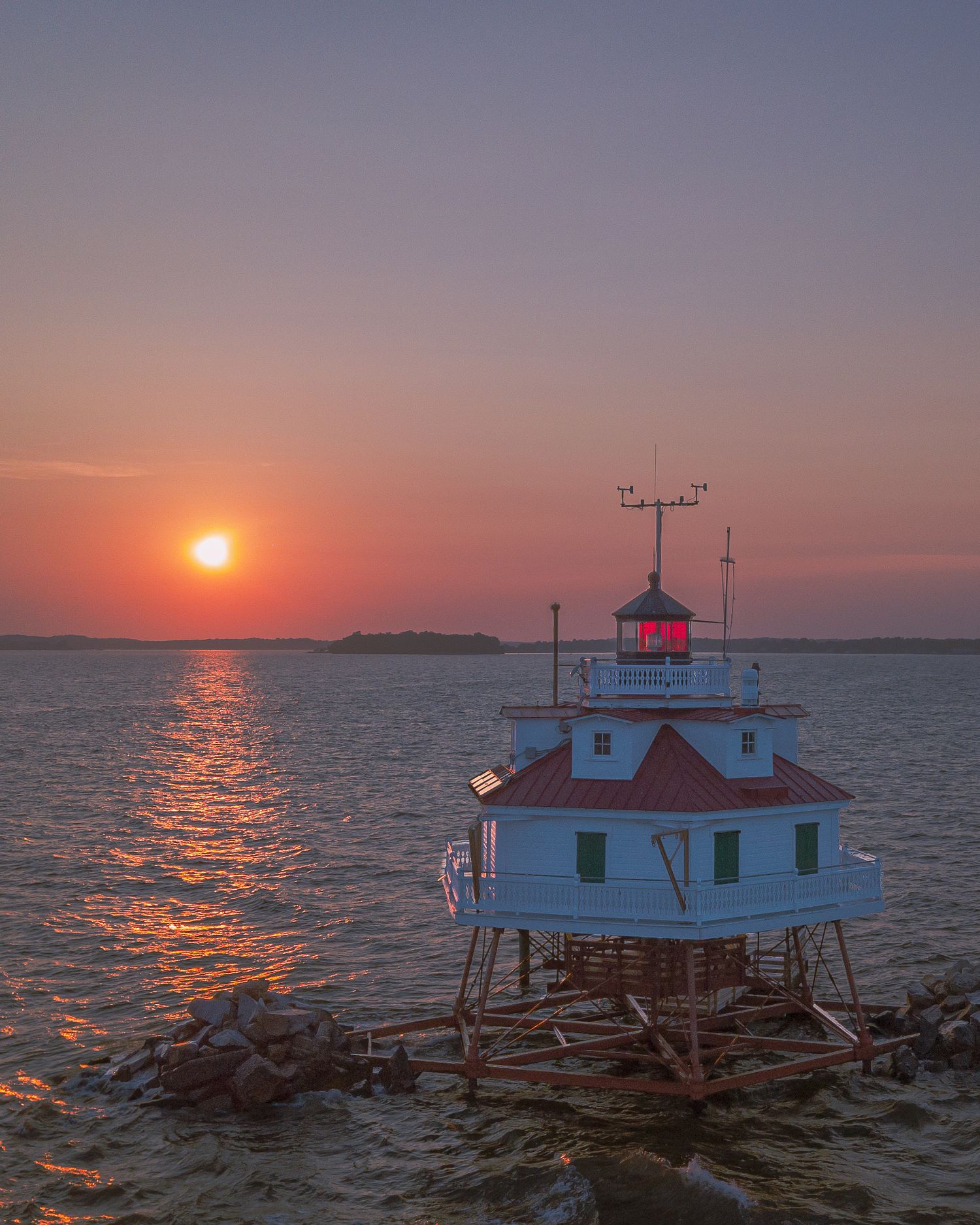 Drone shot of a lighthouse at sunset with warm orange and pink sky tones.