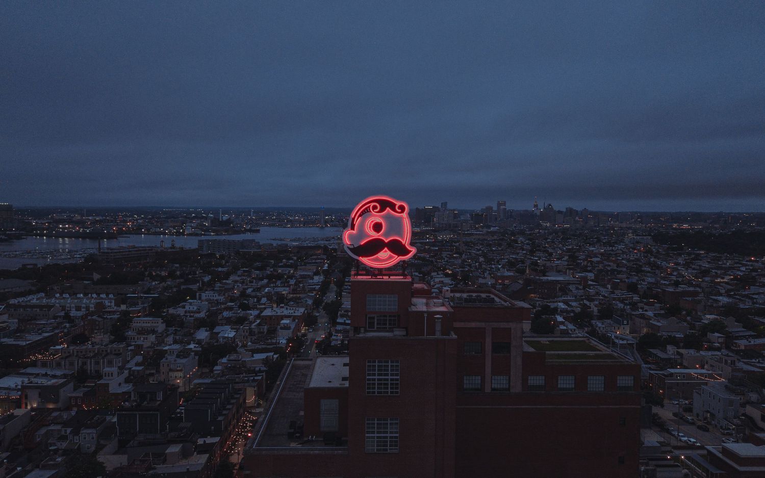 Aerial Baltimore cityscape at dusk featuring a building with illuminated Natty Boh signage against a darkening sky.
