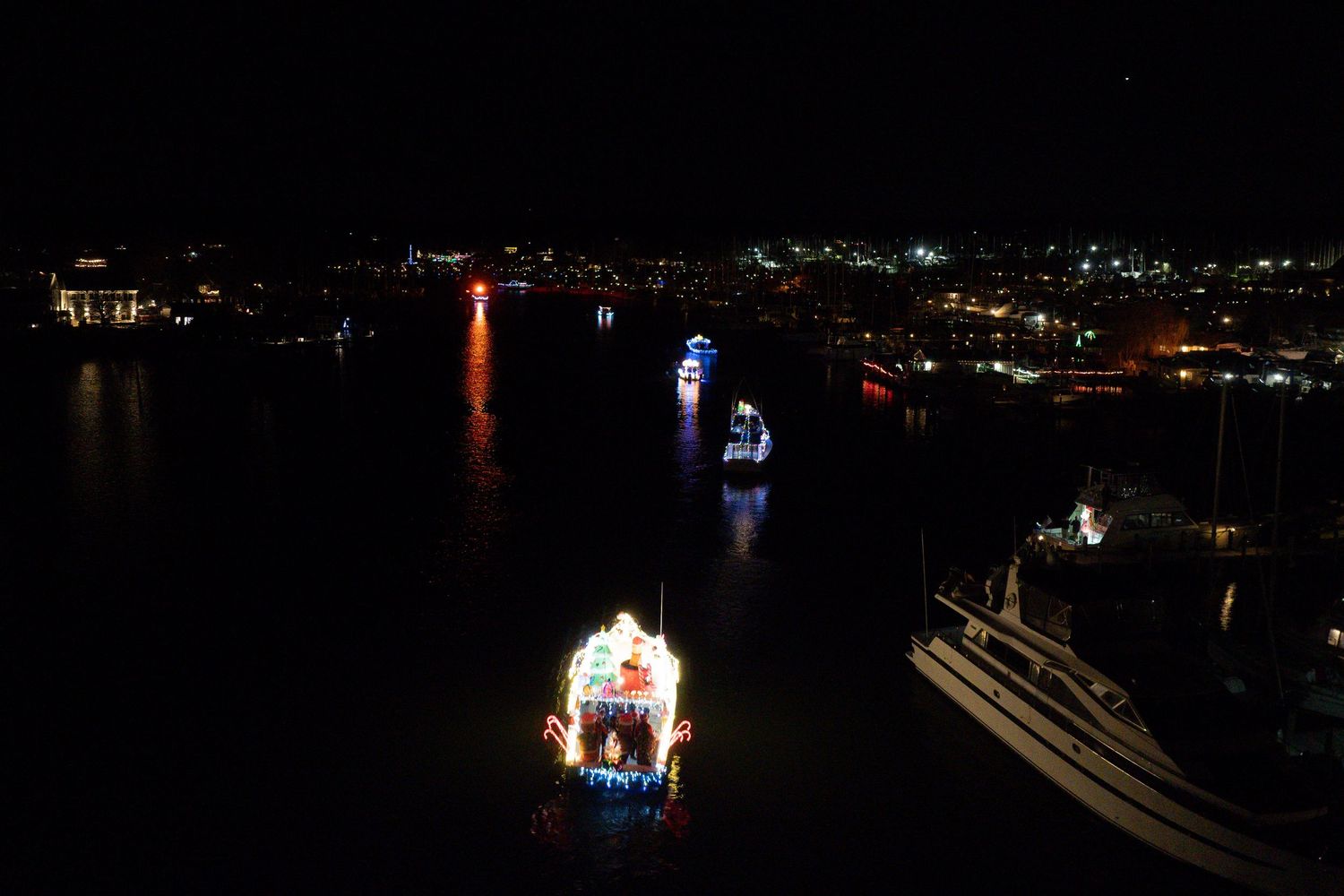Aerial view of multiple boats traveling along a waterway illuminated by city lights.