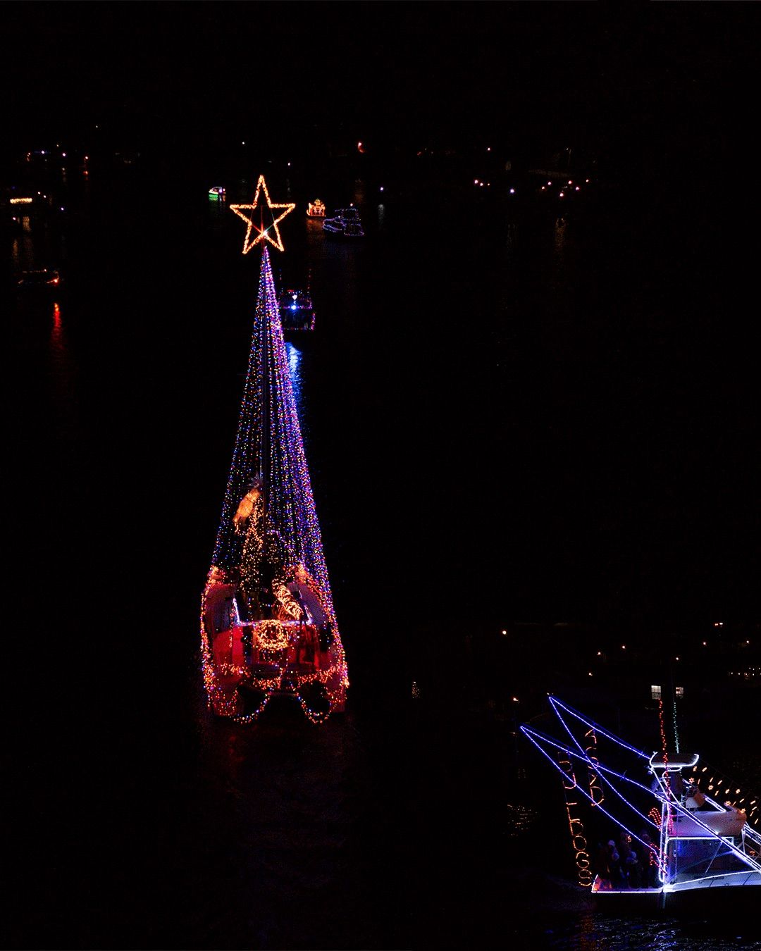 Aerial view of multiple boats traveling along a waterway illuminated by city lights.