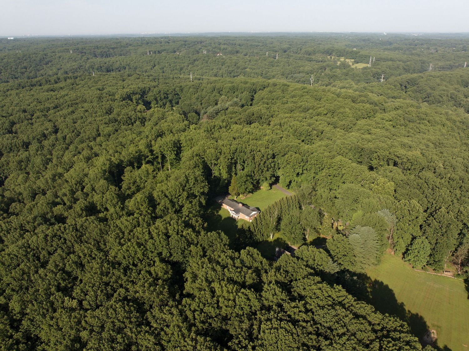 Aerial view of a house for sale set between wooded areas with a highway in the distance.