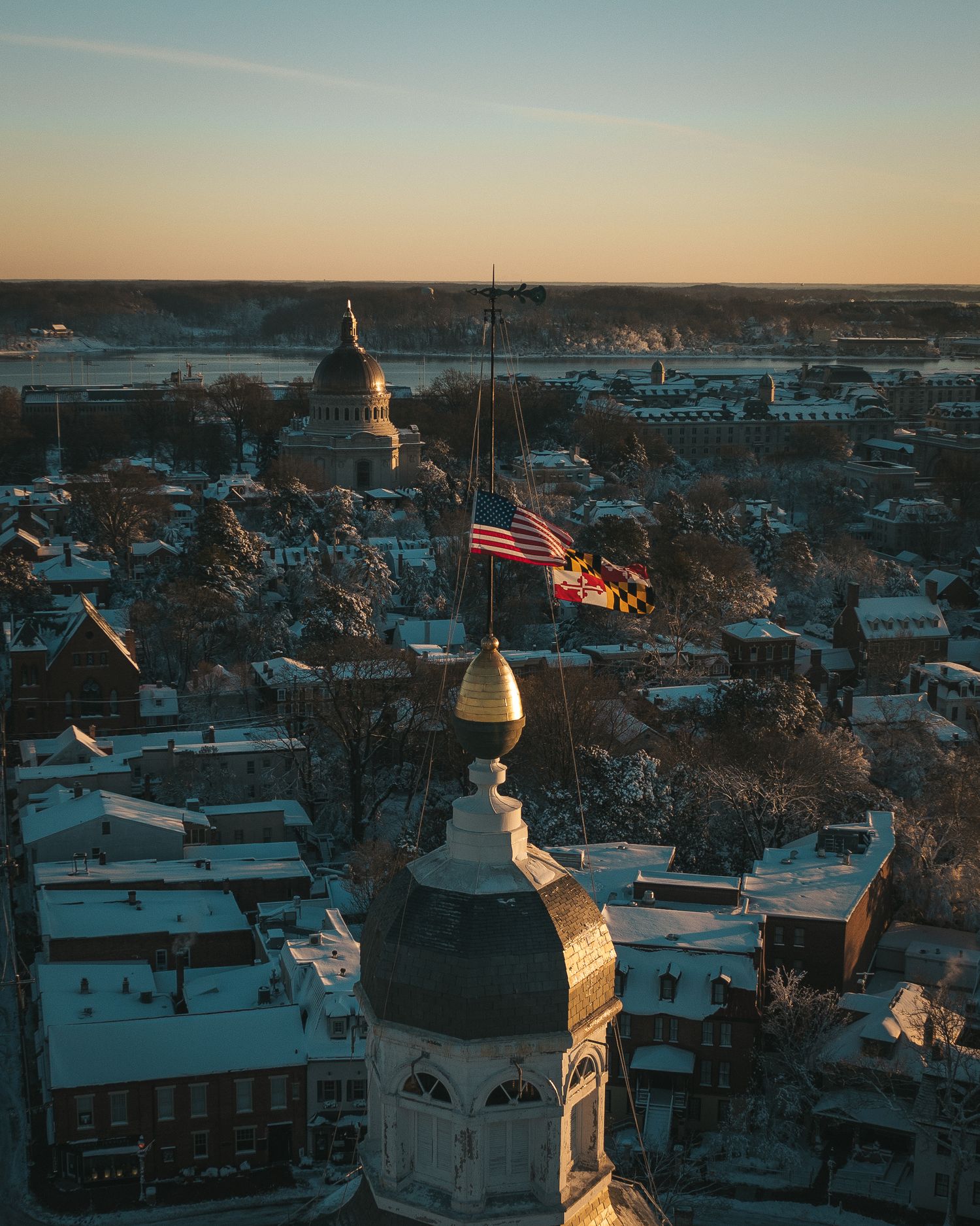 Drone photo of Annapolis Capitol flags at dusk with streets illuminated by evening lights and covered in snow.