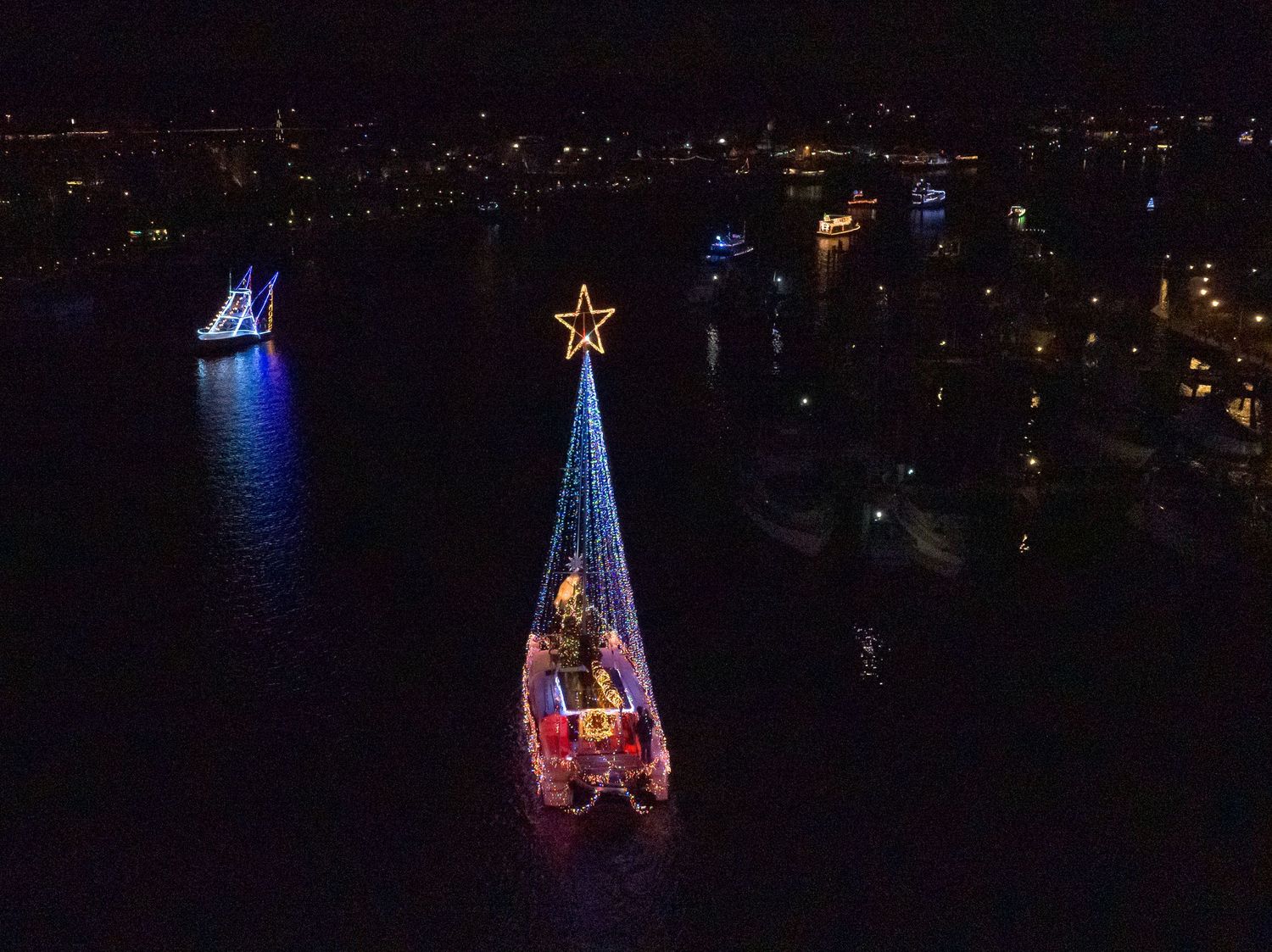 Aerial view of multiple boats traveling along a waterway illuminated by city lights.