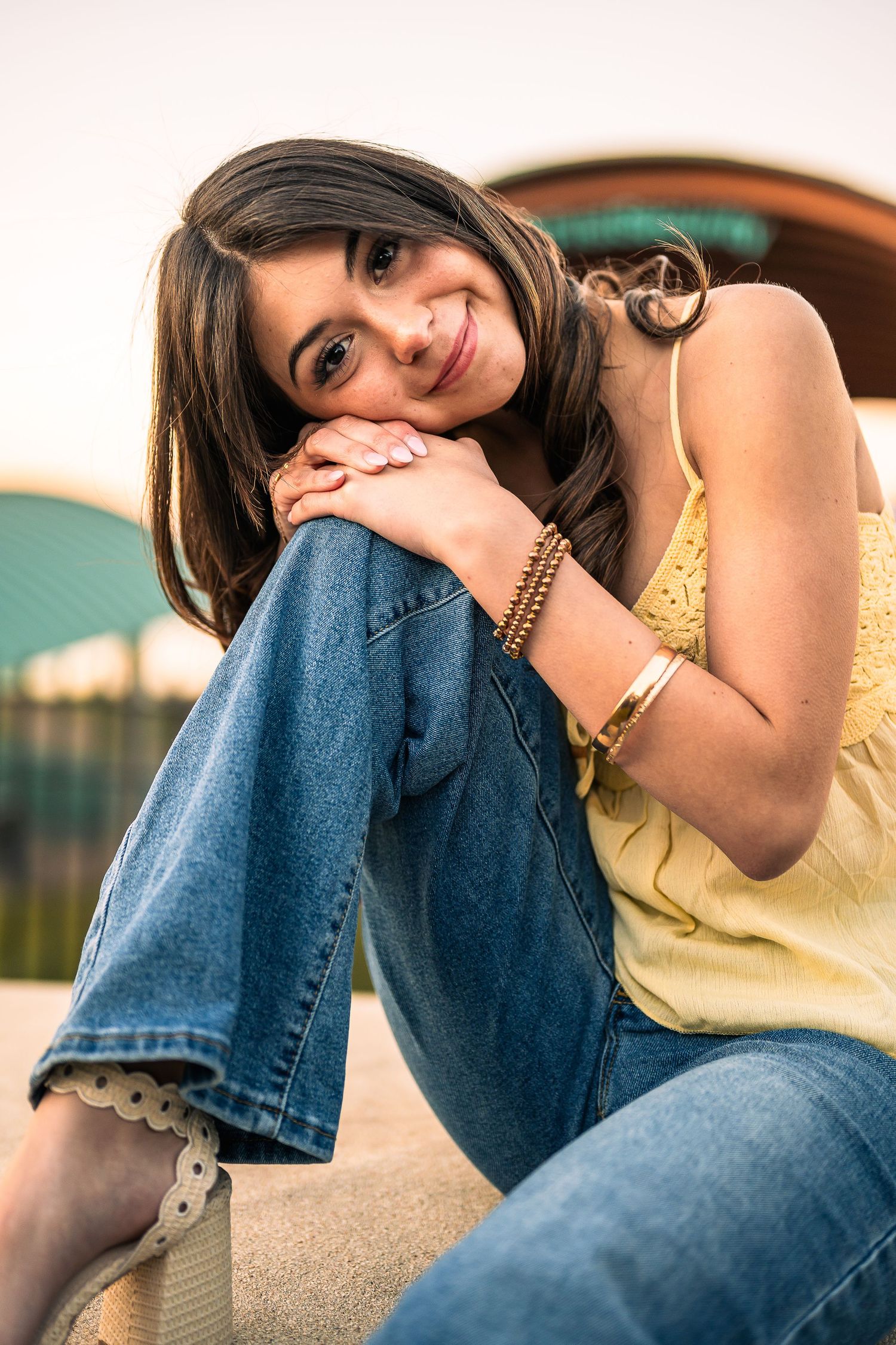Young woman in yellow top and jeans sits outdoors, smiling warmly with hands clasped over knee at sunset.