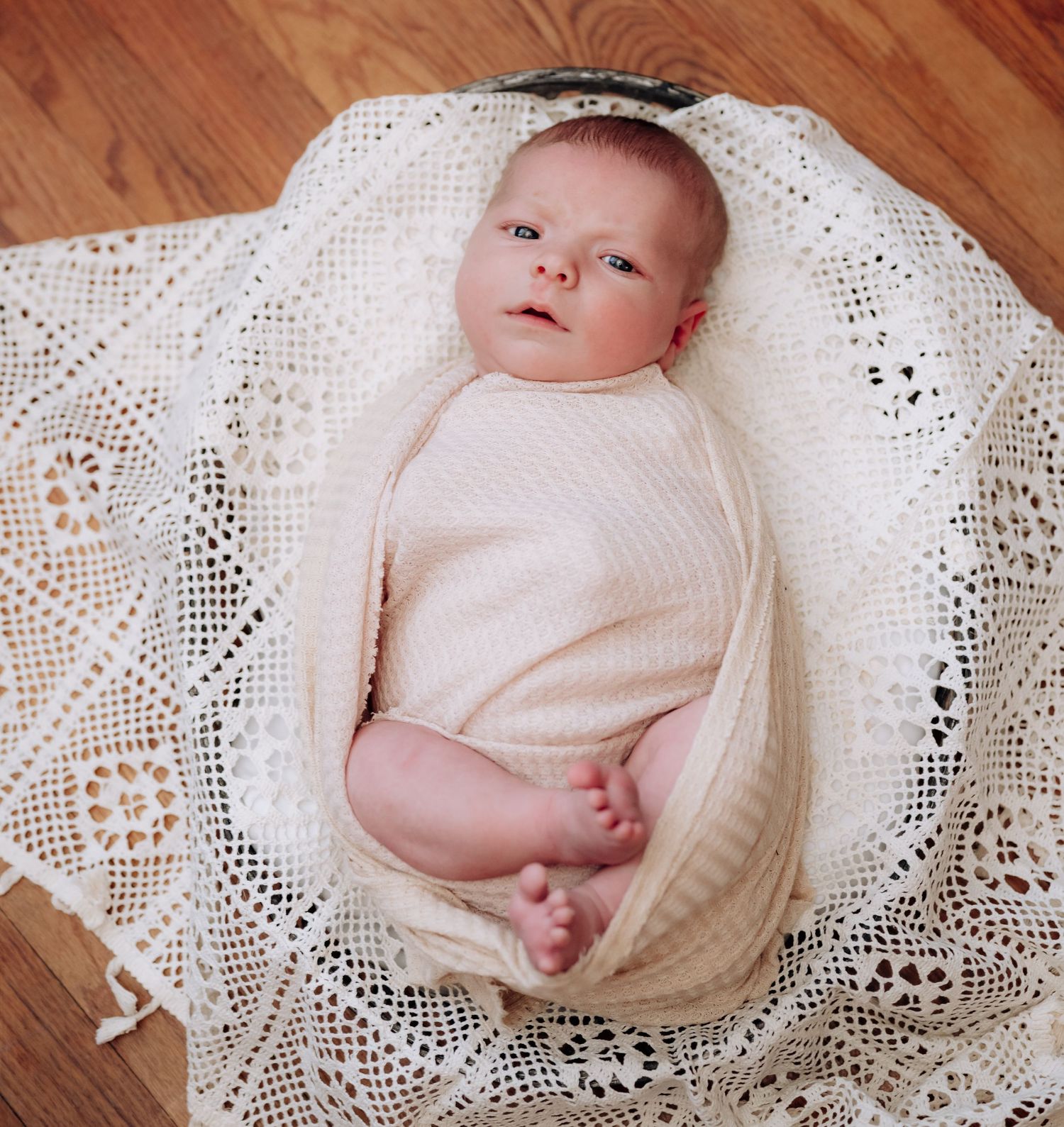 Newborn baby wrapped in soft white lace blanket lying on wooden floor in natural light.
