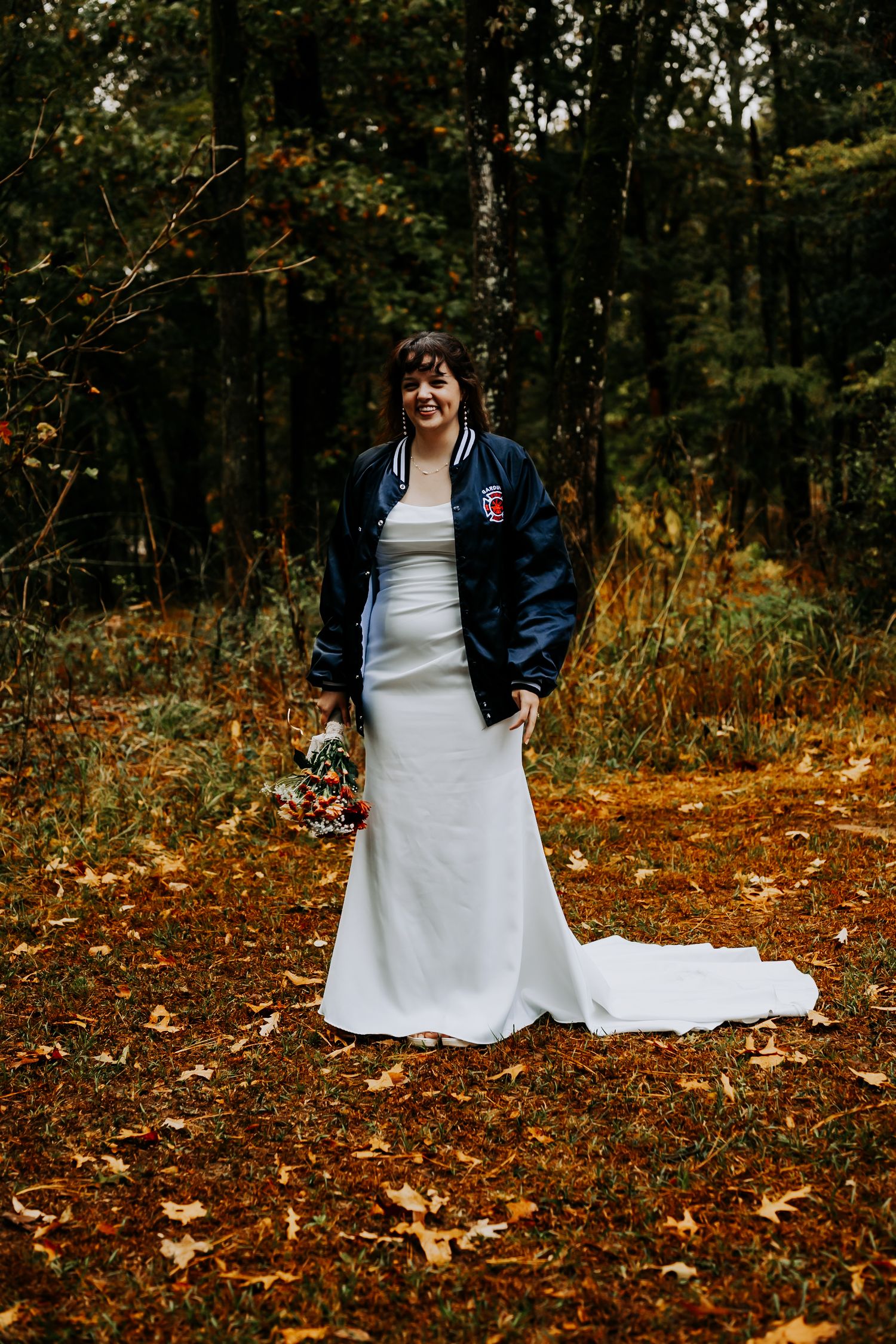 A bride in a white wedding dress poses in autumn woods while wearing a casual blue jacket over their gown.