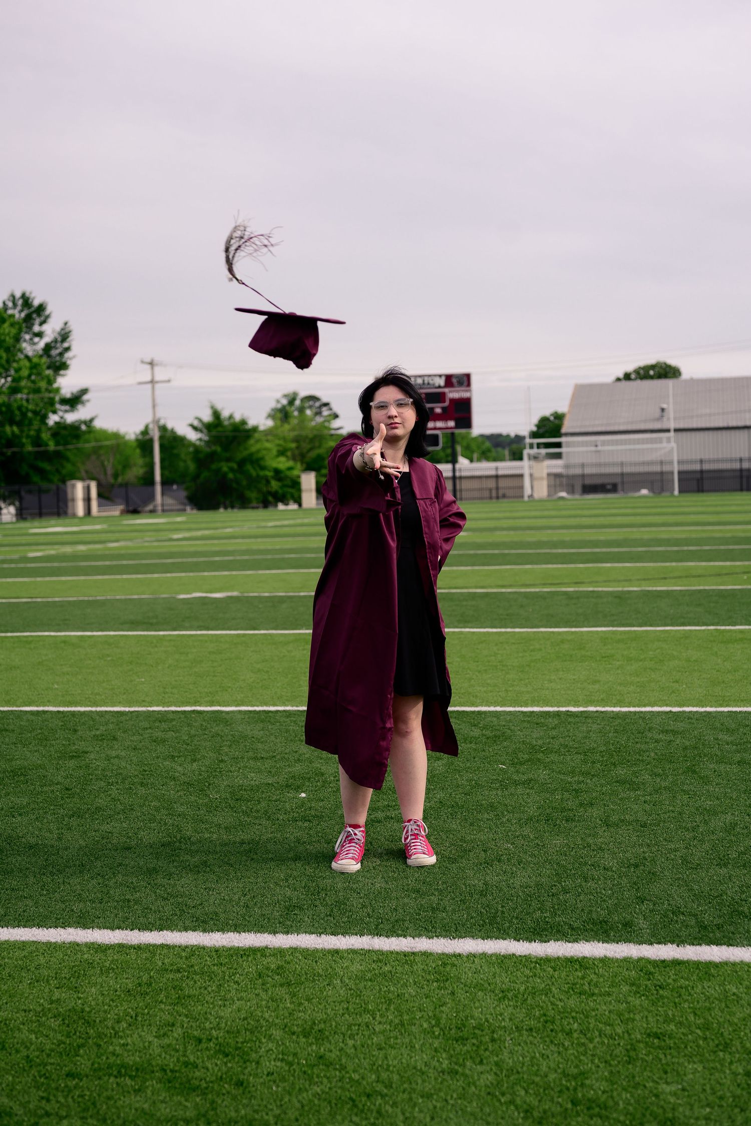 Graduate in maroon cap and gown tossing mortarboard on green football field, celebrating graduation outdoors.
