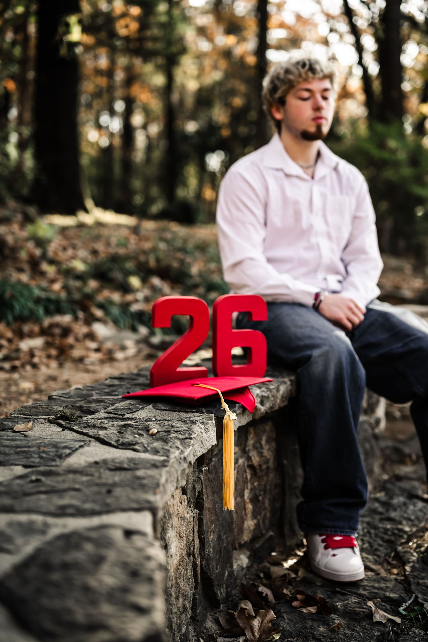 Young man sitting on stone wall in forest with red graduation cap and number 26 in foreground.