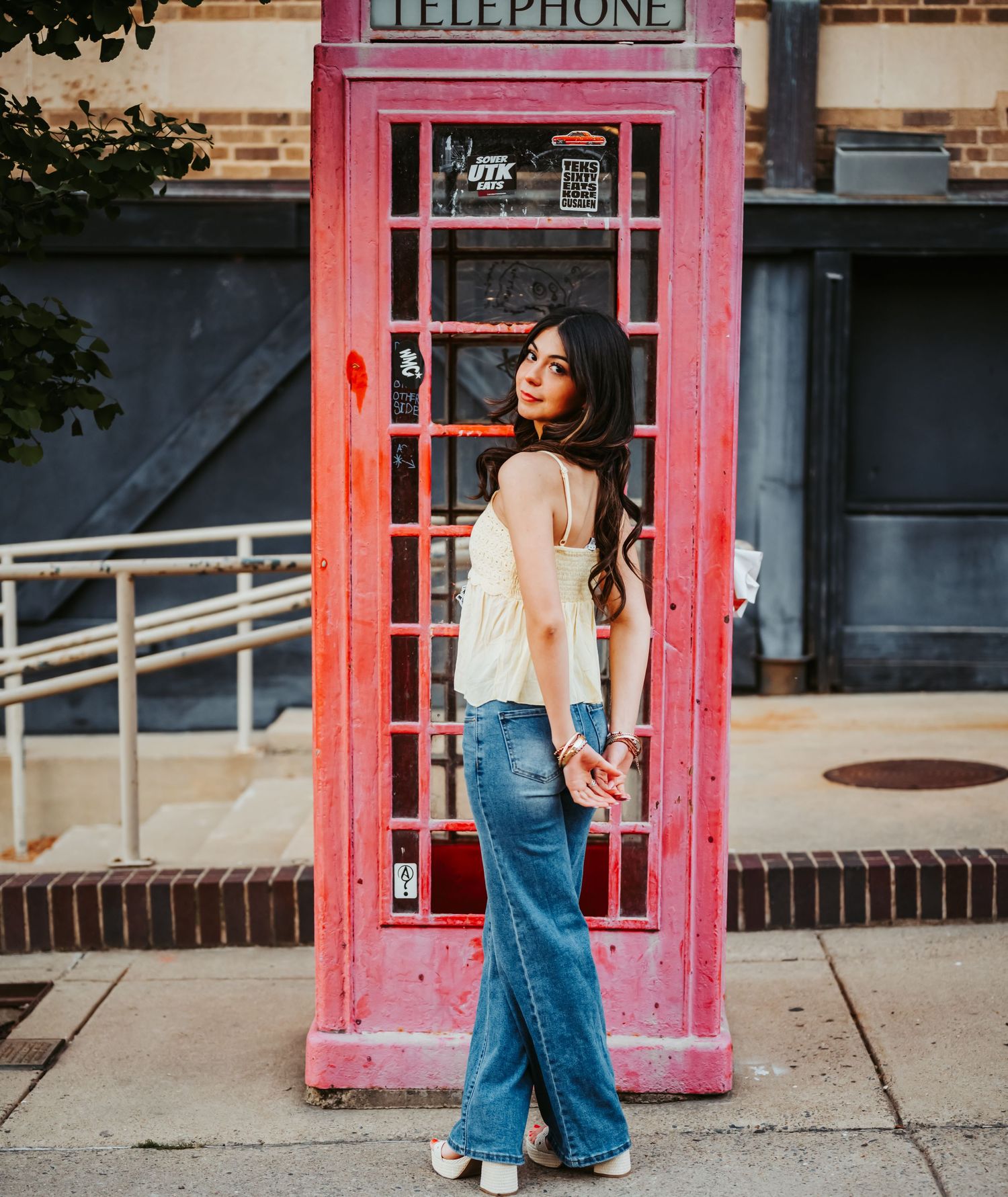 Young woman in flared jeans and white top poses by a red British telephone booth on a city sidewalk.