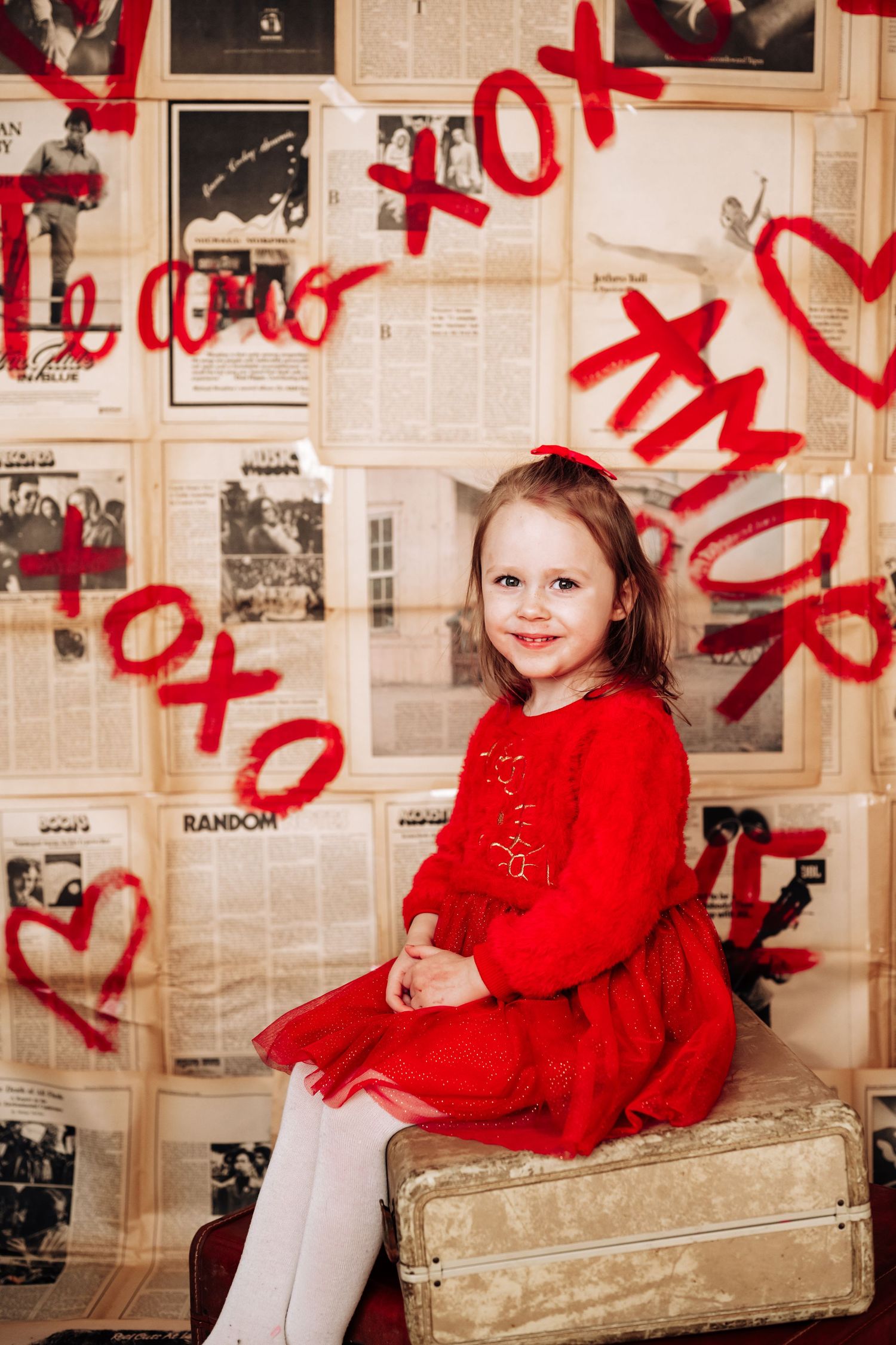 Young girl in red dress sitting on vintage suitcase with XOXO Valentine's Day backdrop.
