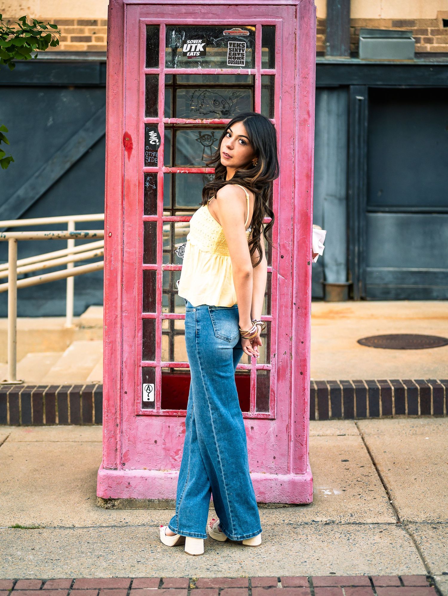 Young woman in yellow top and wide-leg jeans posing by a pink phone booth on a brick sidewalk.