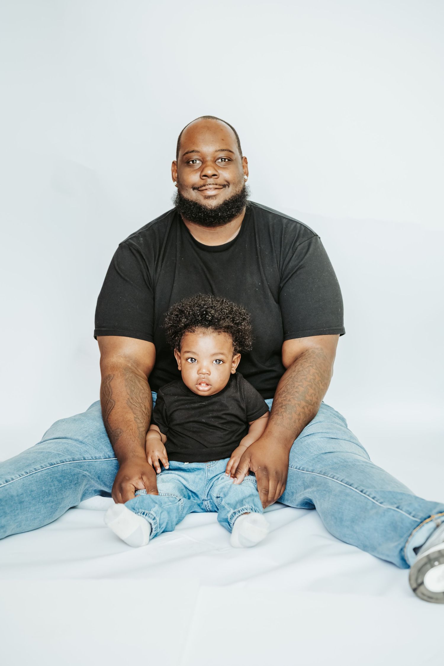 A father and baby boy in matching black shirts and jeans sit together on a white background.