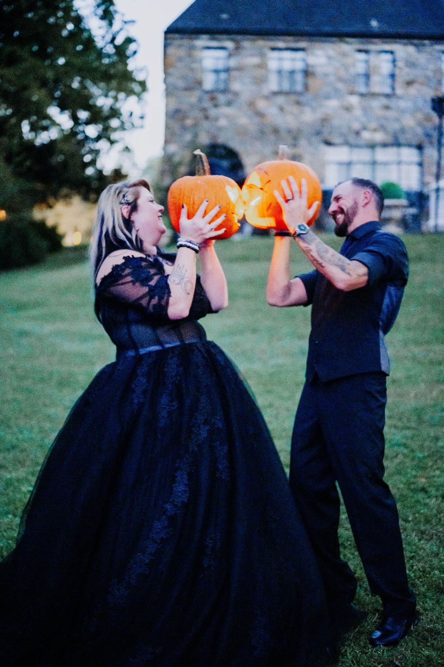 Couple in formal wear holding jack-o'-lanterns in front of a stone building.