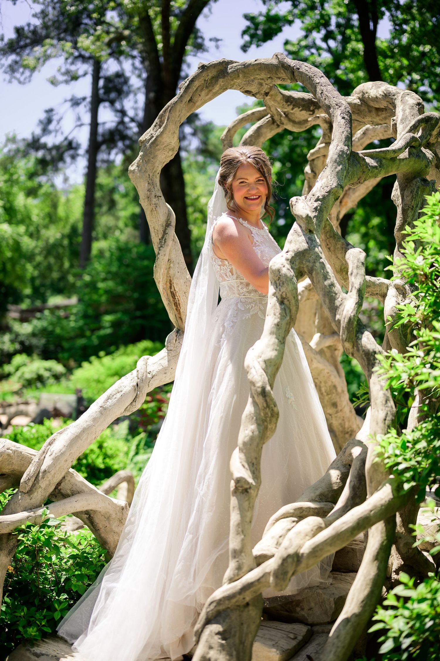 Smiling bride in white lace gown and veil posing under a twisted driftwood arch in a lush green garden.