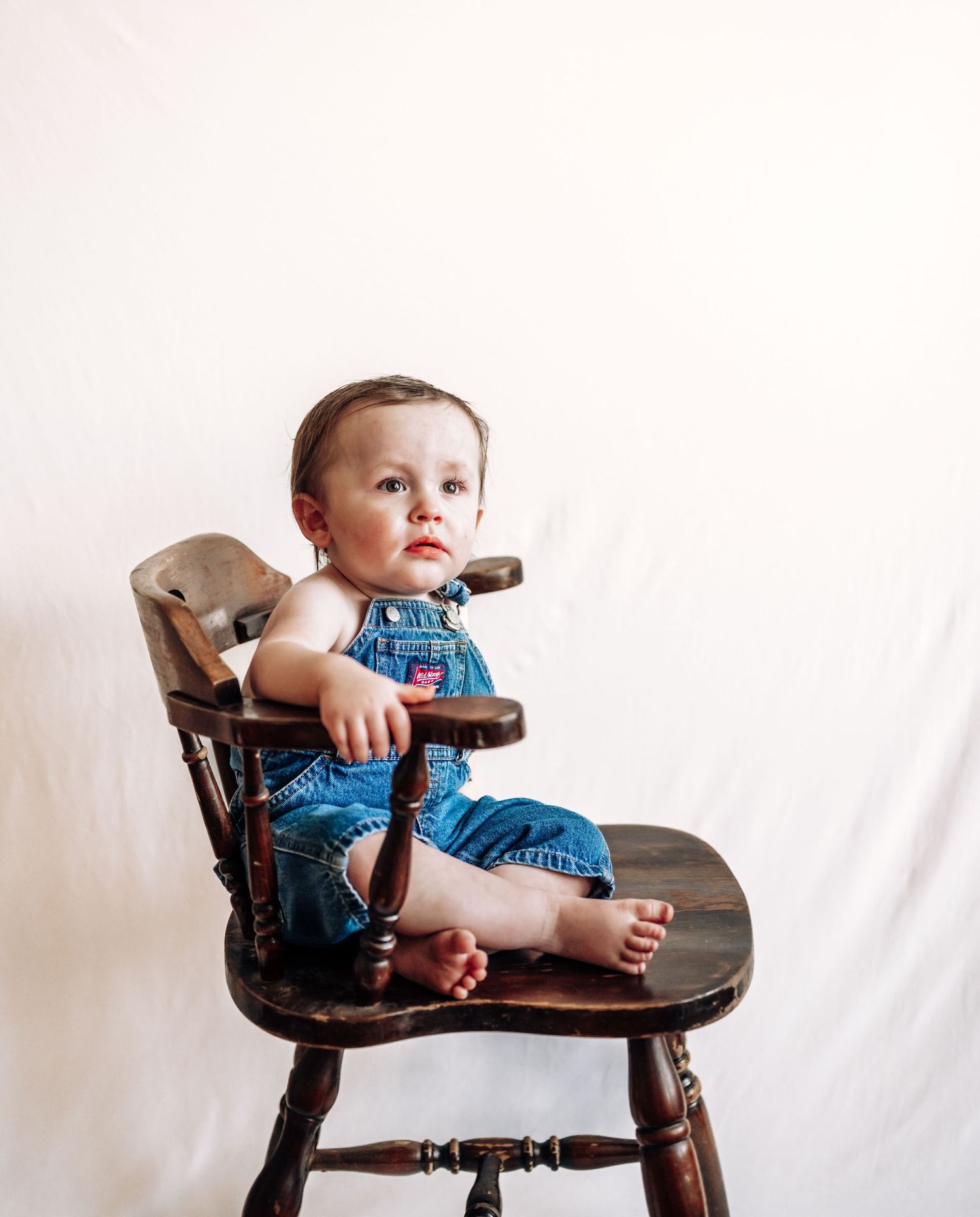 A small child wearing blue overalls sits in a vintage wooden high chair against a white backdrop.